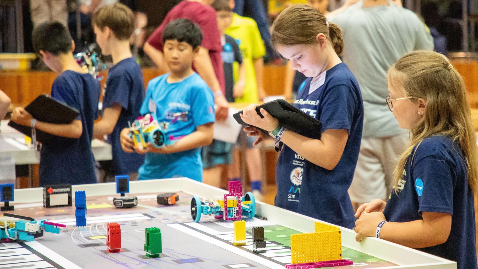 Kinder stehen Tischen mit zusammengesetzten Lego-Bauteilen, einige Kinder halten Tablets in den Händen.