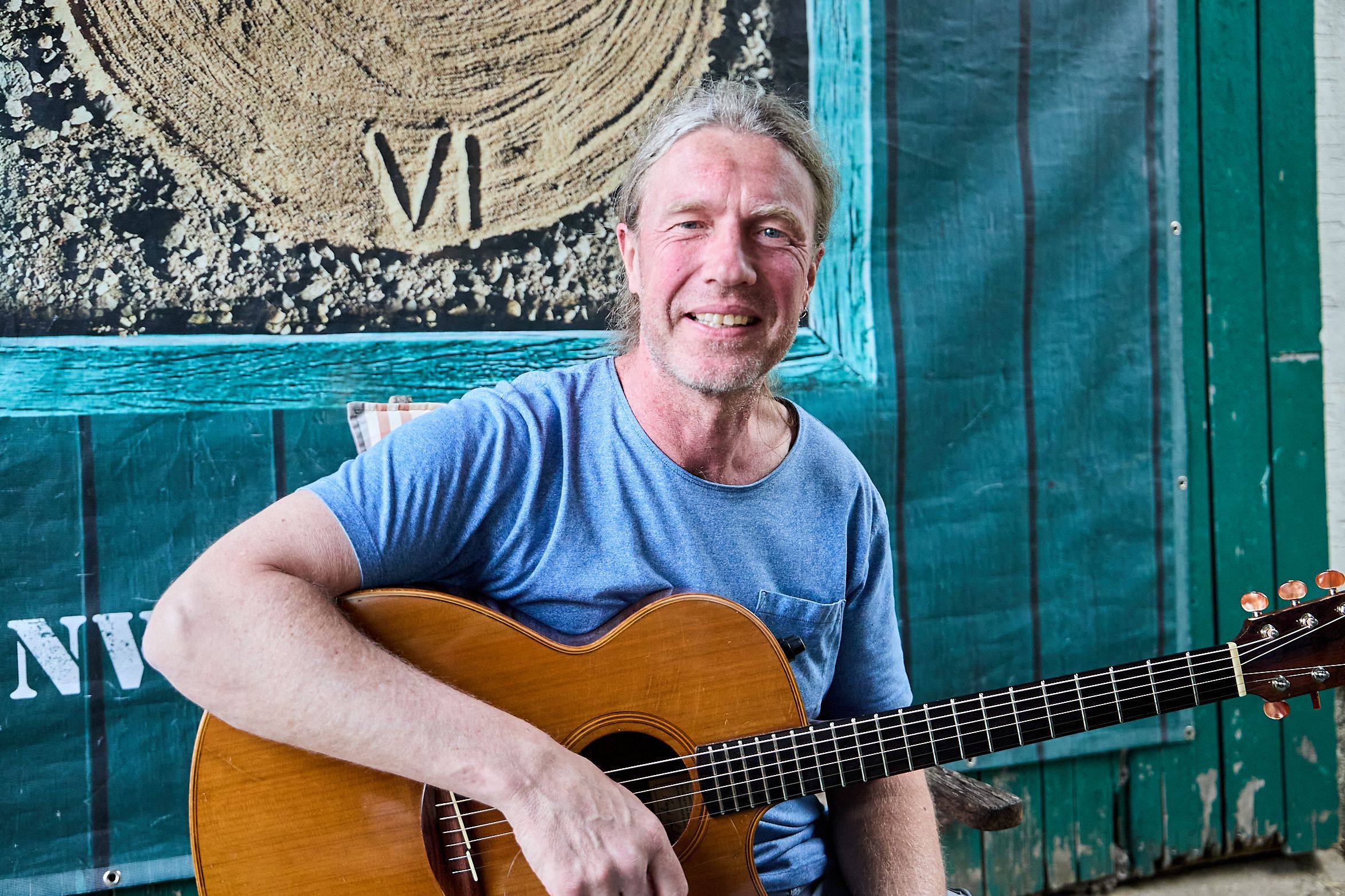 Oliver Kerk hat seine Gitarre in der Hand und sitzt vor einer Holzwand.