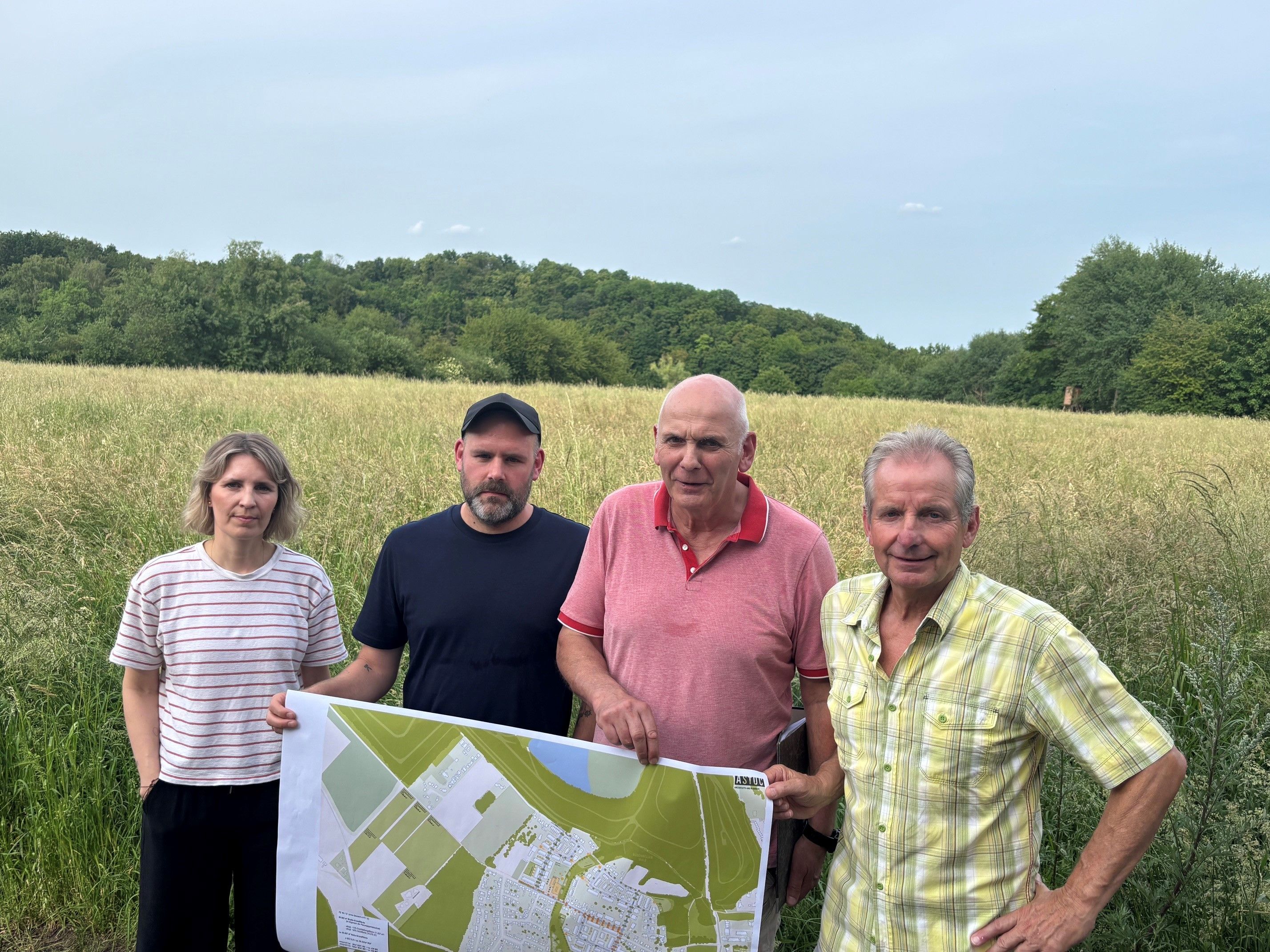 Auf dem Bild ist eine Gruppe von drei Männern und einer Frau zu sehen, die vor einem Feld stehen. Sie halten einen Plan in der Hand.