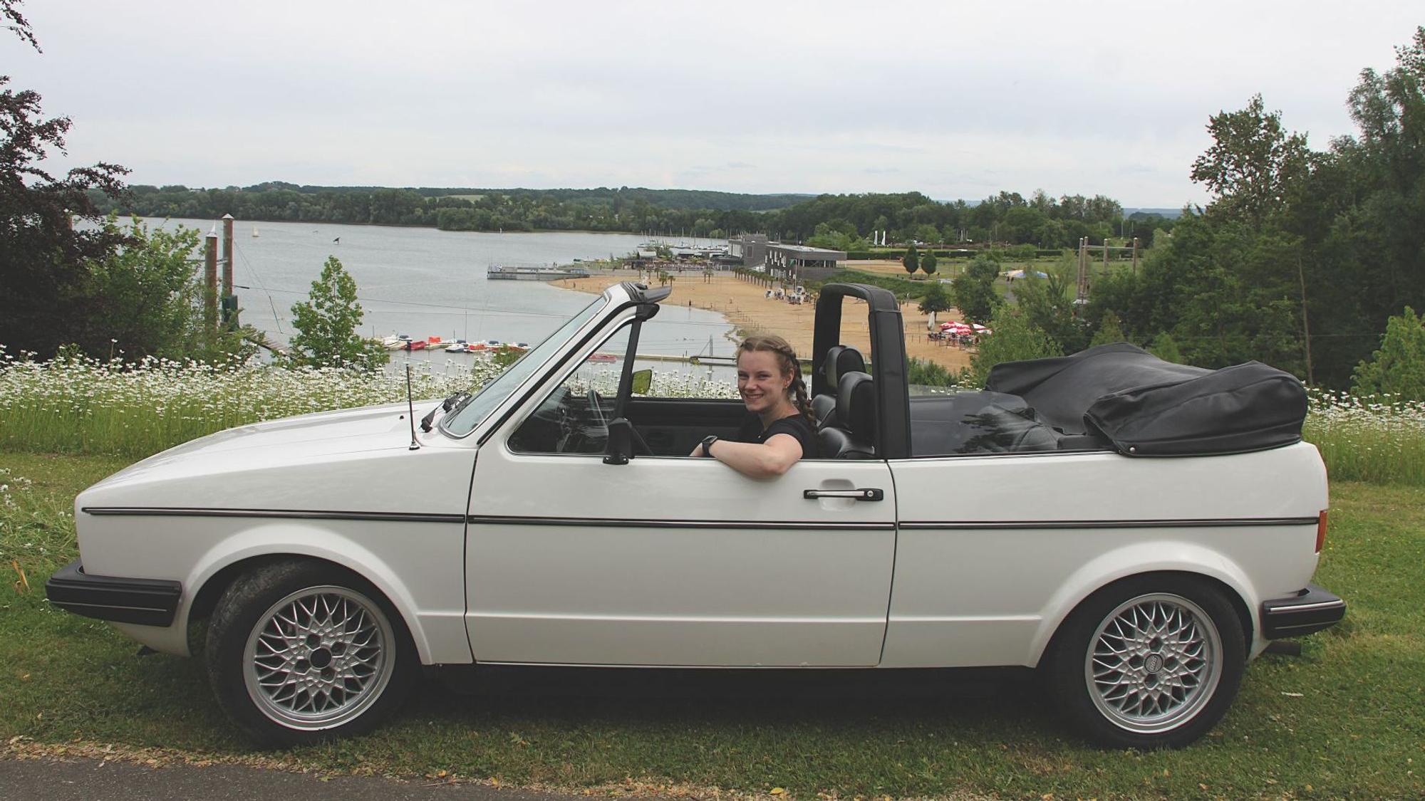In einem weißen Golf-Cabriolet sitzt eine junge Frau. Im Hintergrund ist der Zülpicher Wassersportsee zu sehen.