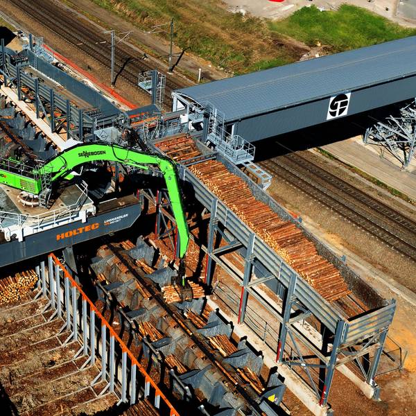 Ein Portalkran bewegt sich in einem Schienensystem mit Brücke über den Holzplatz.