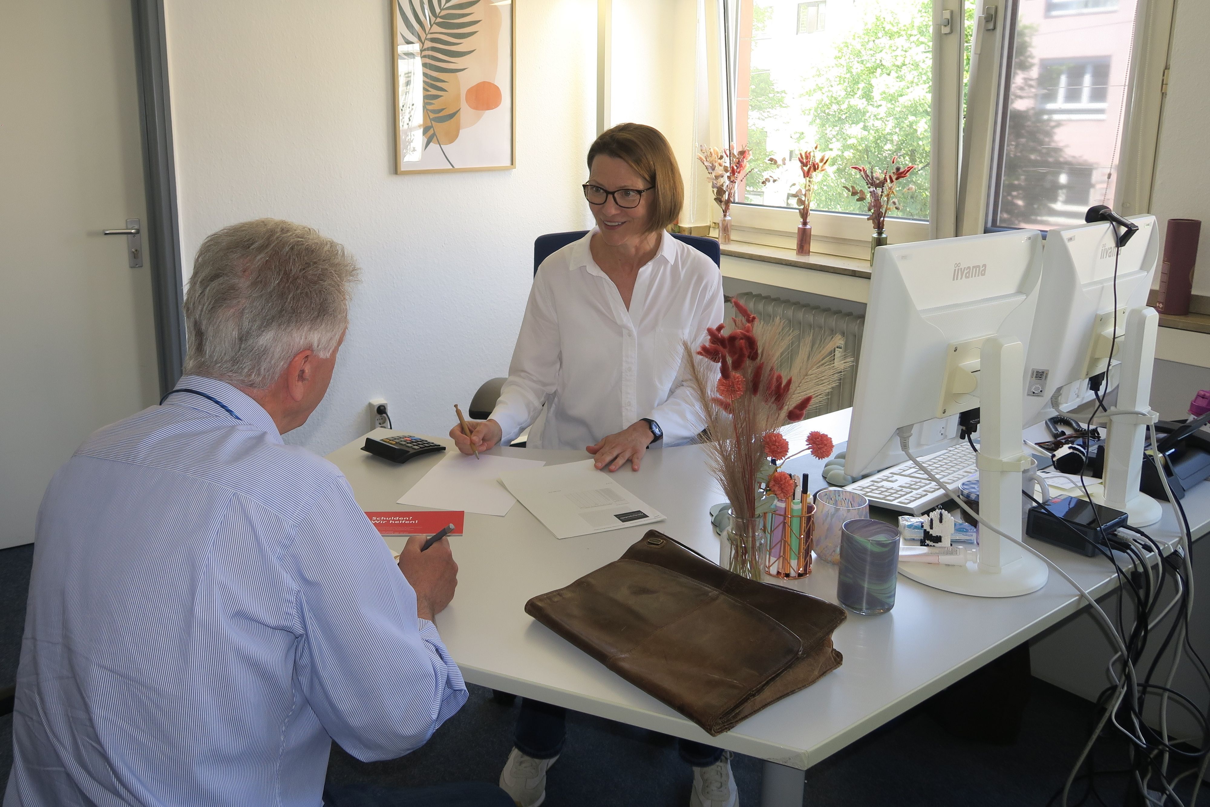 Eine Frau sitzt hinter einem Schreibtisch einem Mann gegenüber. Beide machen sich Notizen.