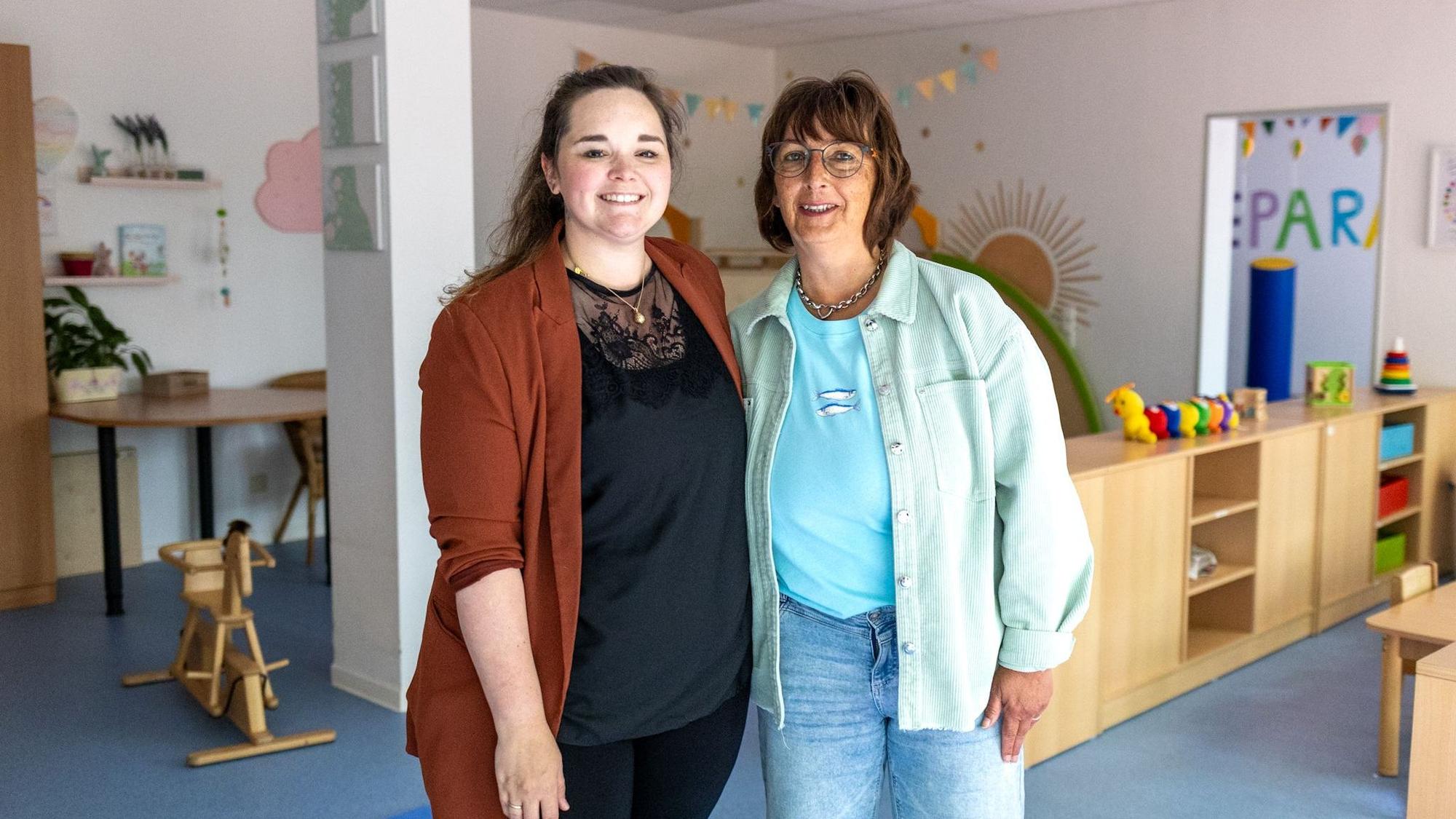 Zwei Frauen stehen Arm in Arm im Spielzimmer und lächeln in die Kamera.