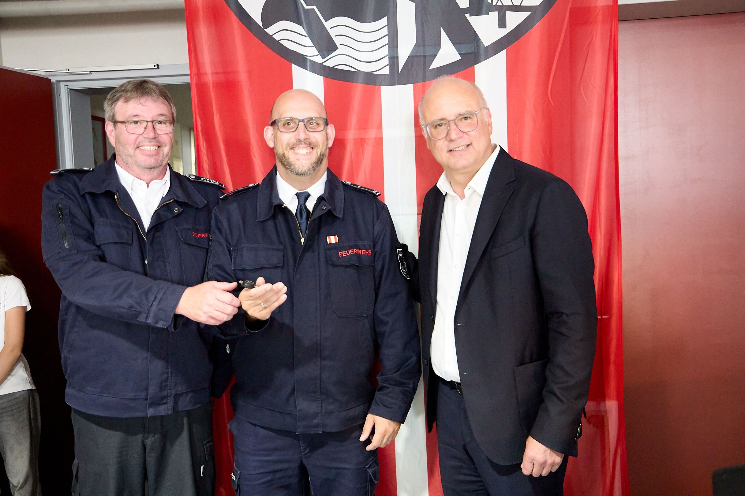 Die drei Männer stehen vor einer Fahne der Feuerwehr.