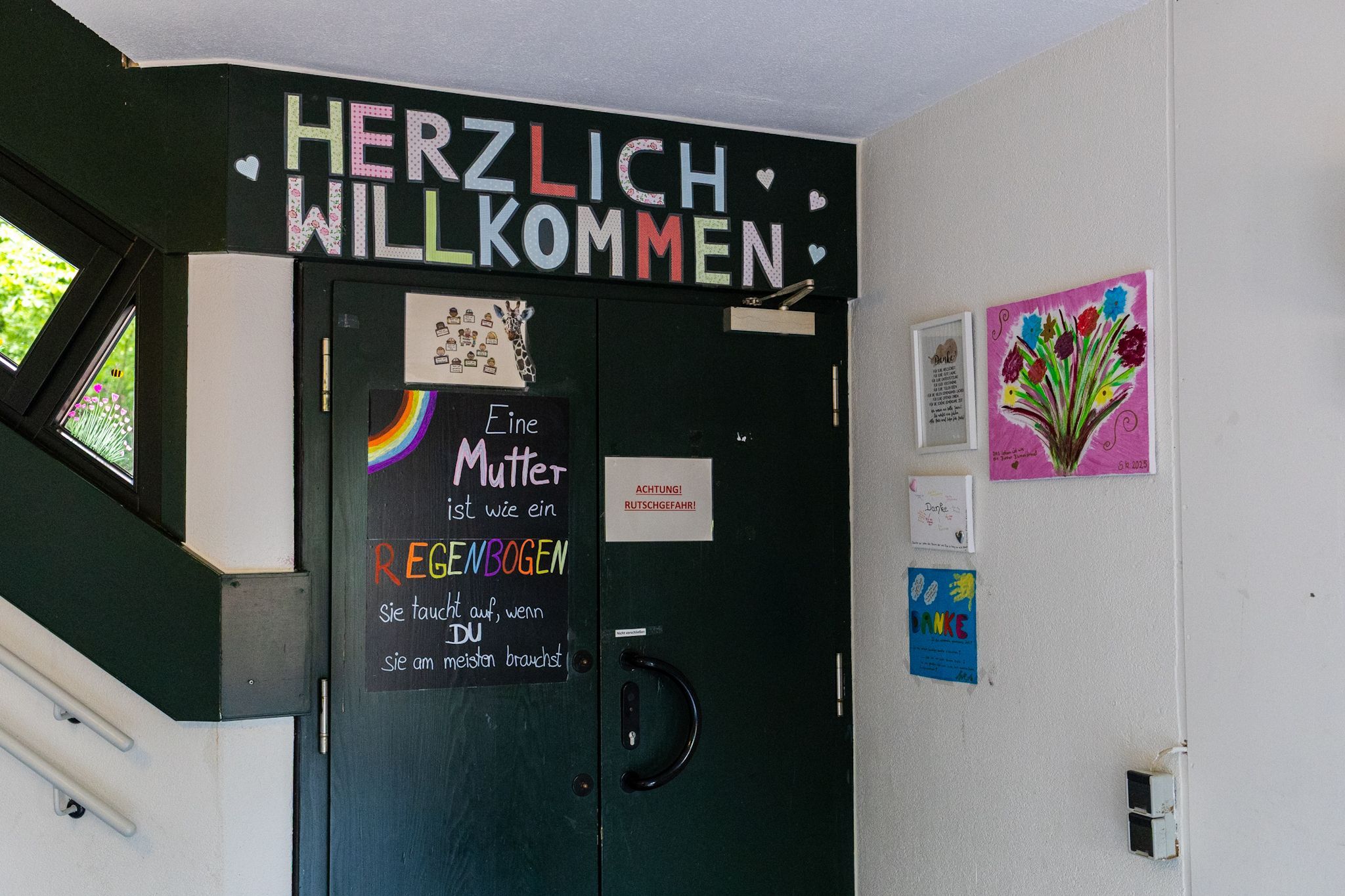 Tür mit „Herzlich Willkommen“, daneben hängen Bilder mit Blumen.