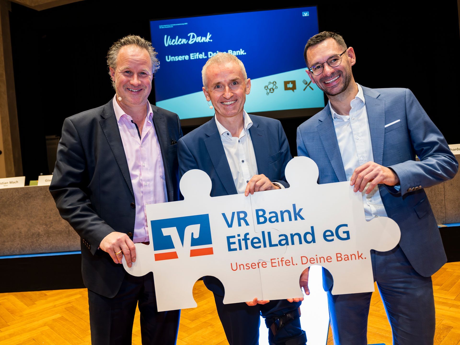 Die drei Bankvorstände halten gemeinsam ein Schild mit dem Logo der Volksbanken, dem Namen „VR Bank EifelLand eG“ und dem Slogan „Unsere Eifel. Deine Bank.““