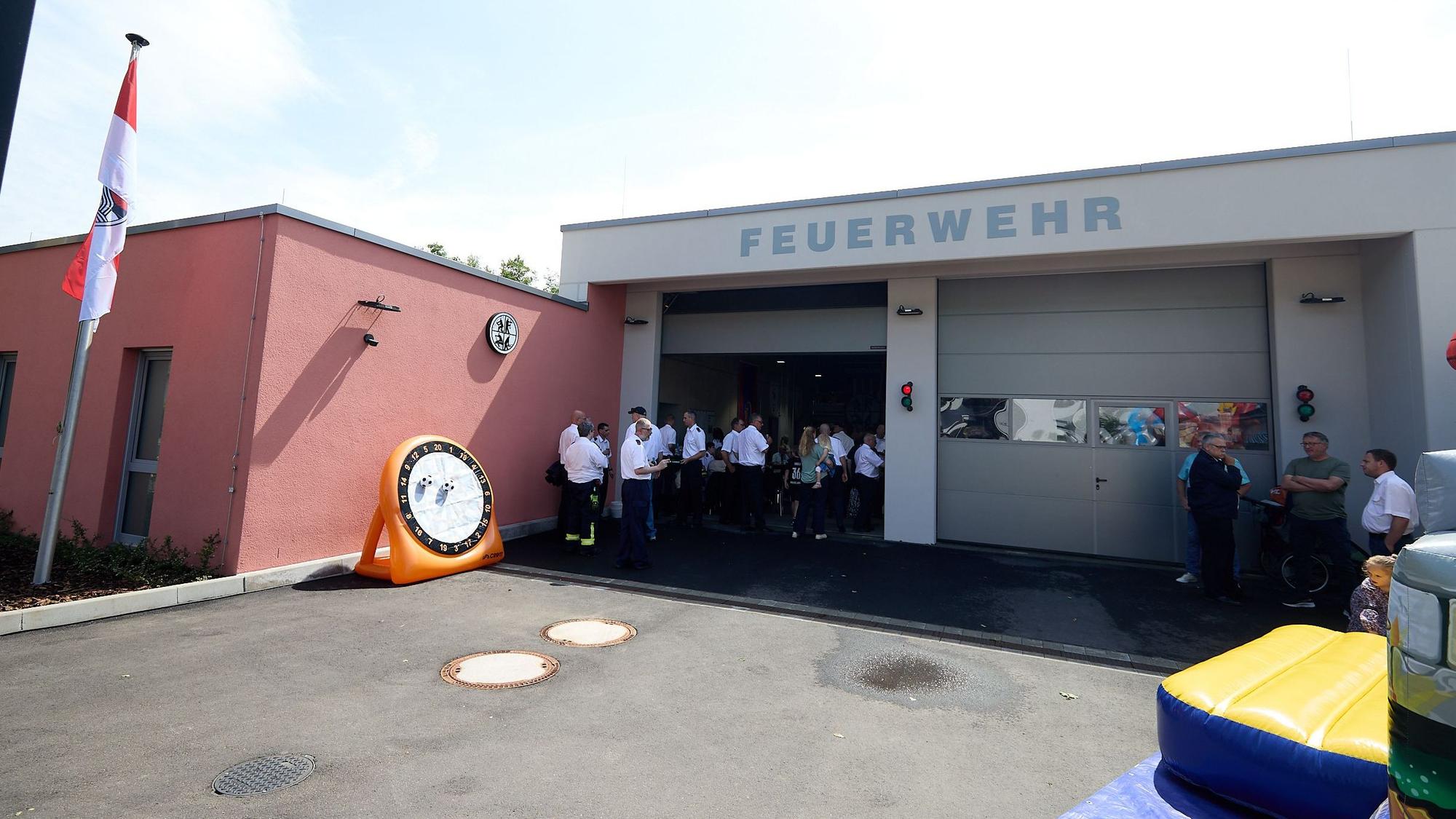 Menschen stehen neben einem roten Gebäude vor weißen Garagen, auf denen in großen Buchstaben Feuerwehr steht.