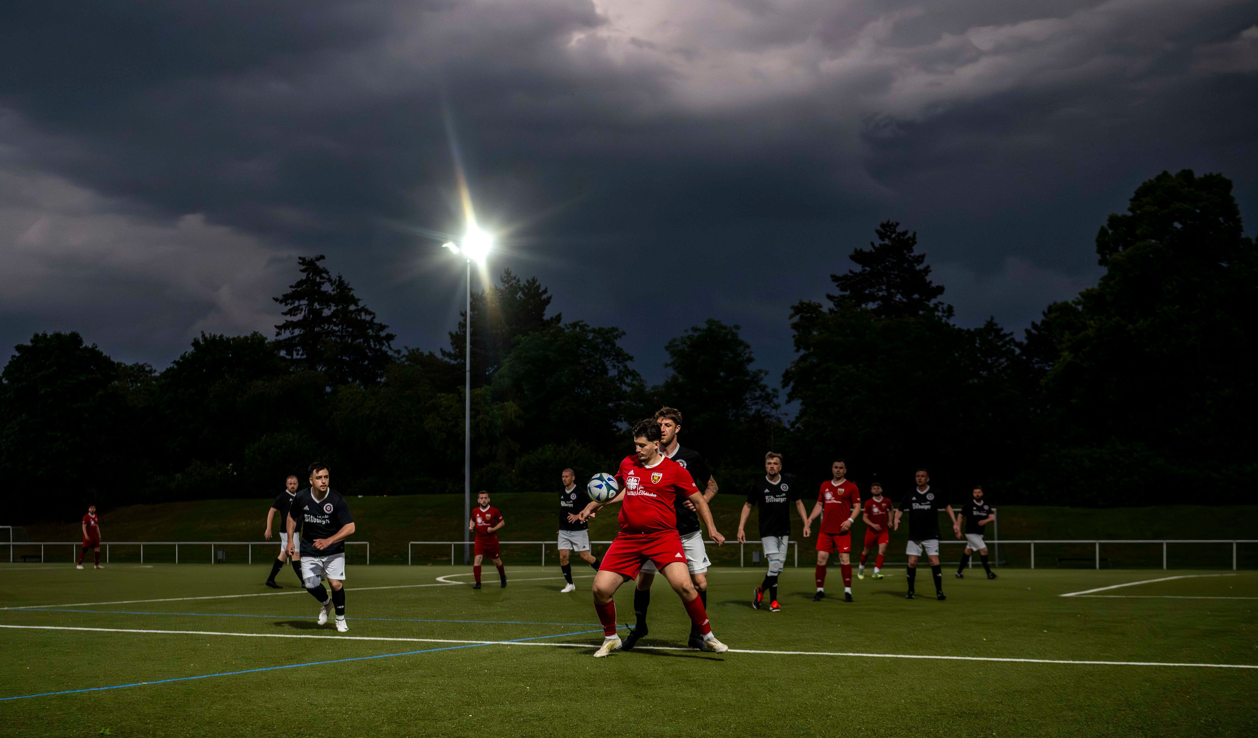 Gewitterwolken tauchen einen Platz in ein dunkles Licht, ein Fußballer in rotem Trikot nimmt einen Ball mit der Brust an.