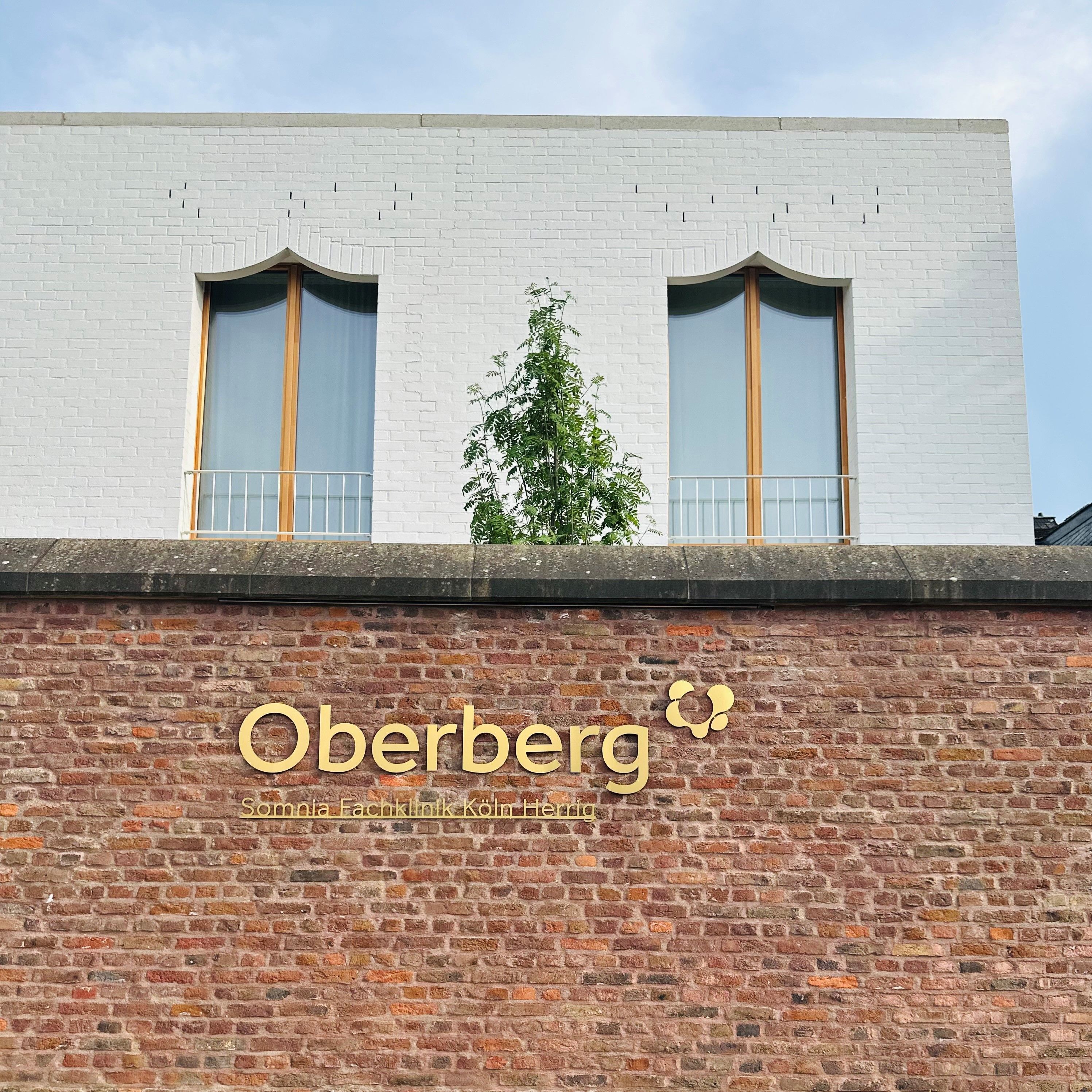 Zu sehen ist eine Mauer vor einem Klinikgebäude mit einem goldenen Schriftzug „Oberberg“.
