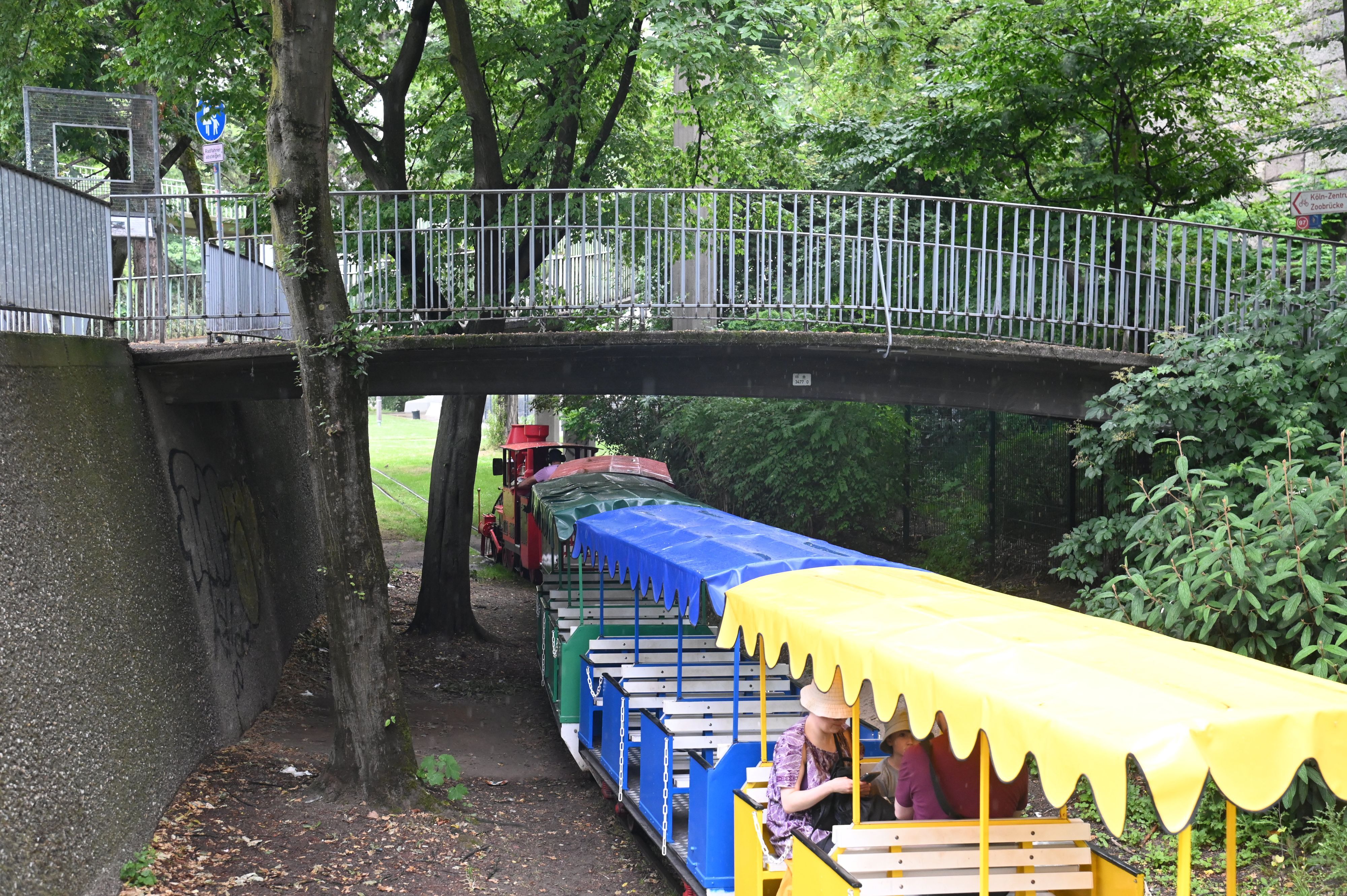 02.06.2026 Köln. Im Rheinpark führt eine kleine Brücke an der Fußgängerampe zur Zoobrücke über die Gleise der dortigen Kleinbahn, die hier ihre Haltestelle „Rosengarten“ hat. Die Parkeisenbahn wurde zur Bundesgartenschau 1971 errichtet. Foto: Alexander Schwaiger
