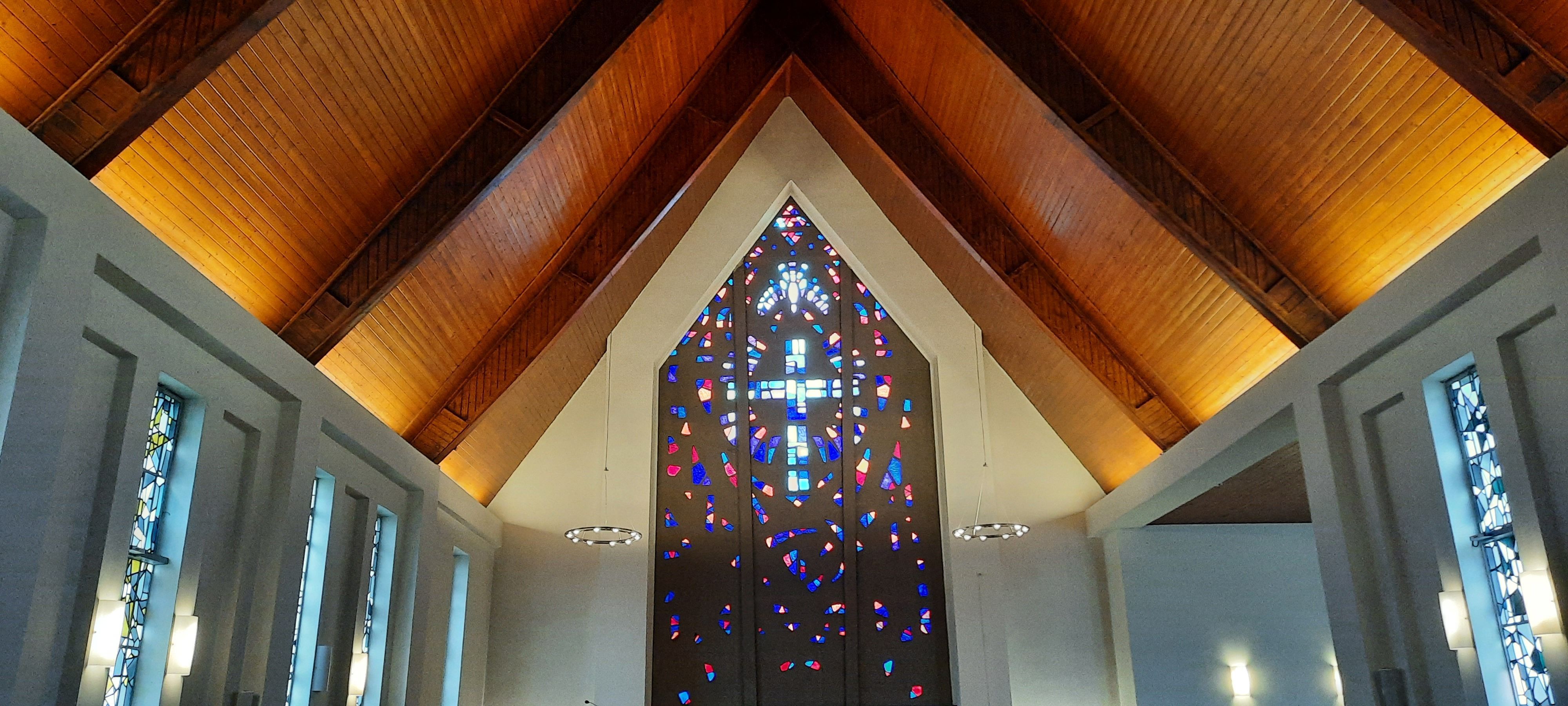 Das Bild zeigt den Innenraum der Kirche. Vorne ist ein Kreuz als buntes Fenstermosaik zu sehen.
