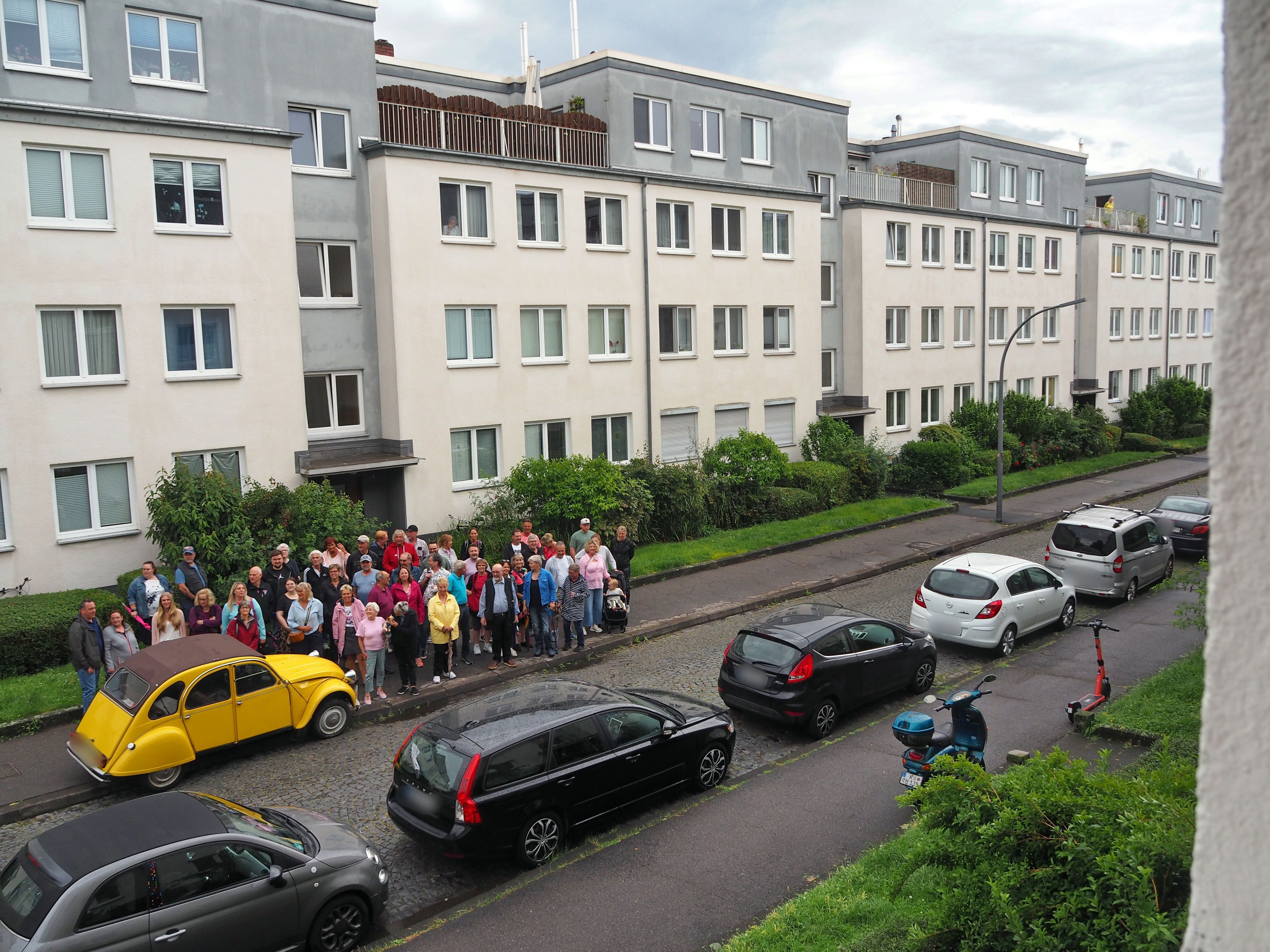 Autos parken an einer Straße, eine Menschengruppe steht auf einer Seite auf dem Bürgersteig.