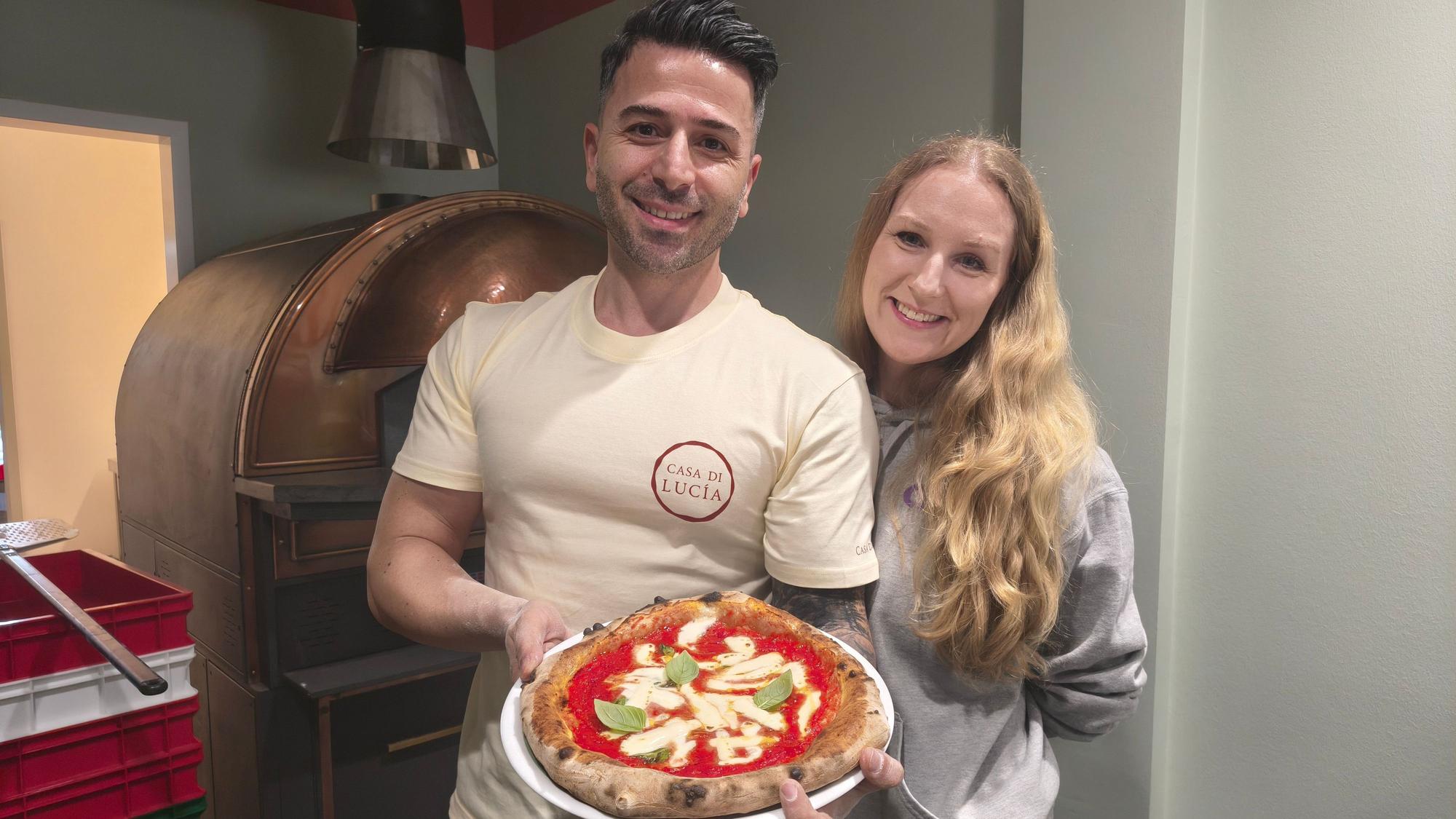 Sinan Batur steht dicht neben seiner Ehefrau Alruna Batur vor einem Ofen und präsentiert eine neapolitanische Pizza auf einem Teller.