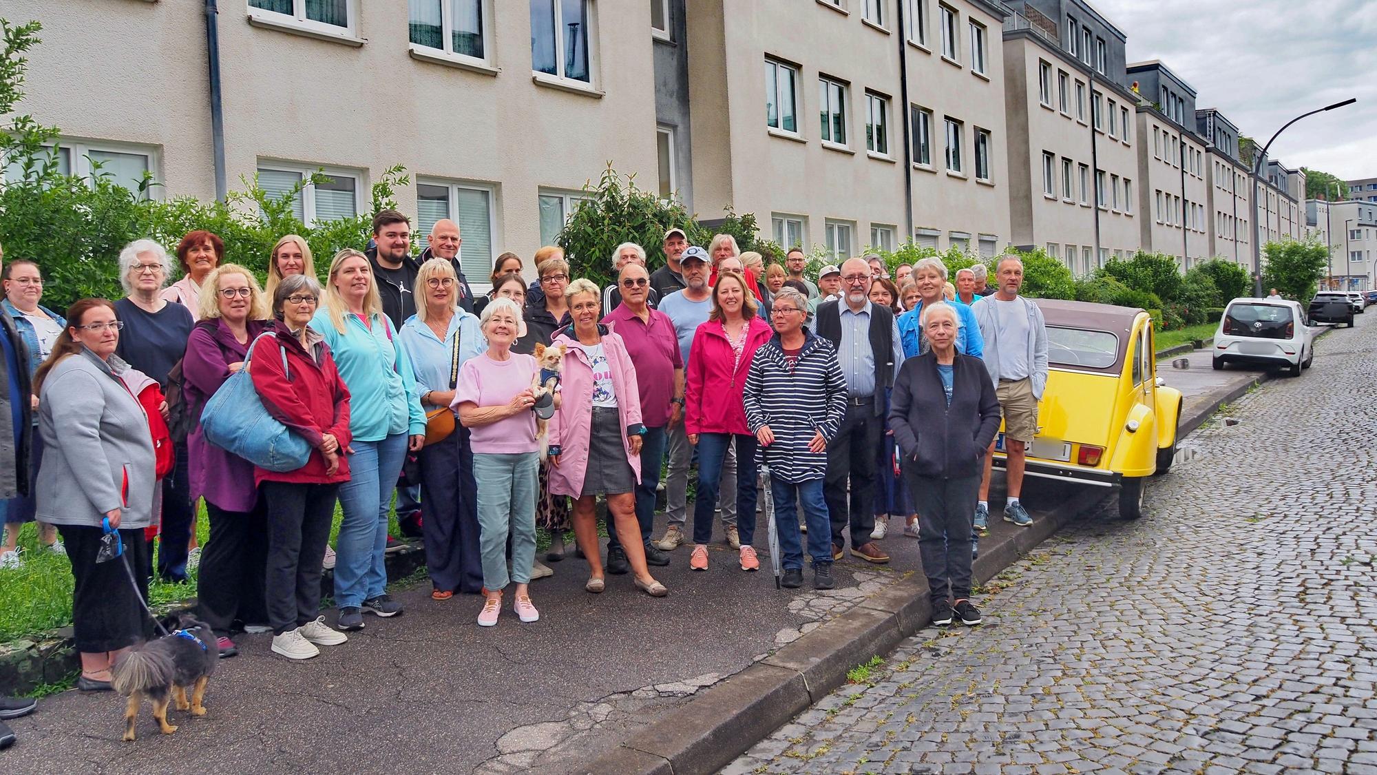 Eine Gruppe Menschen steht auf einem Gehweg, eine Ente parkt auf dem Bürgersteig.