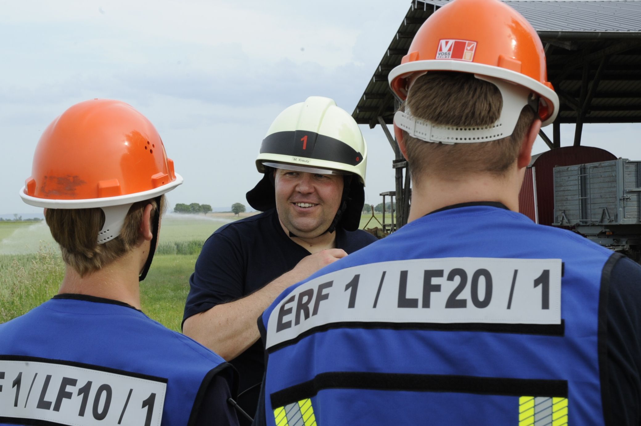 Ein Mann mit Helm spricht mit zwei anderen jungen Männern in orangenen Helmen, die von hinten zu sehen sind.