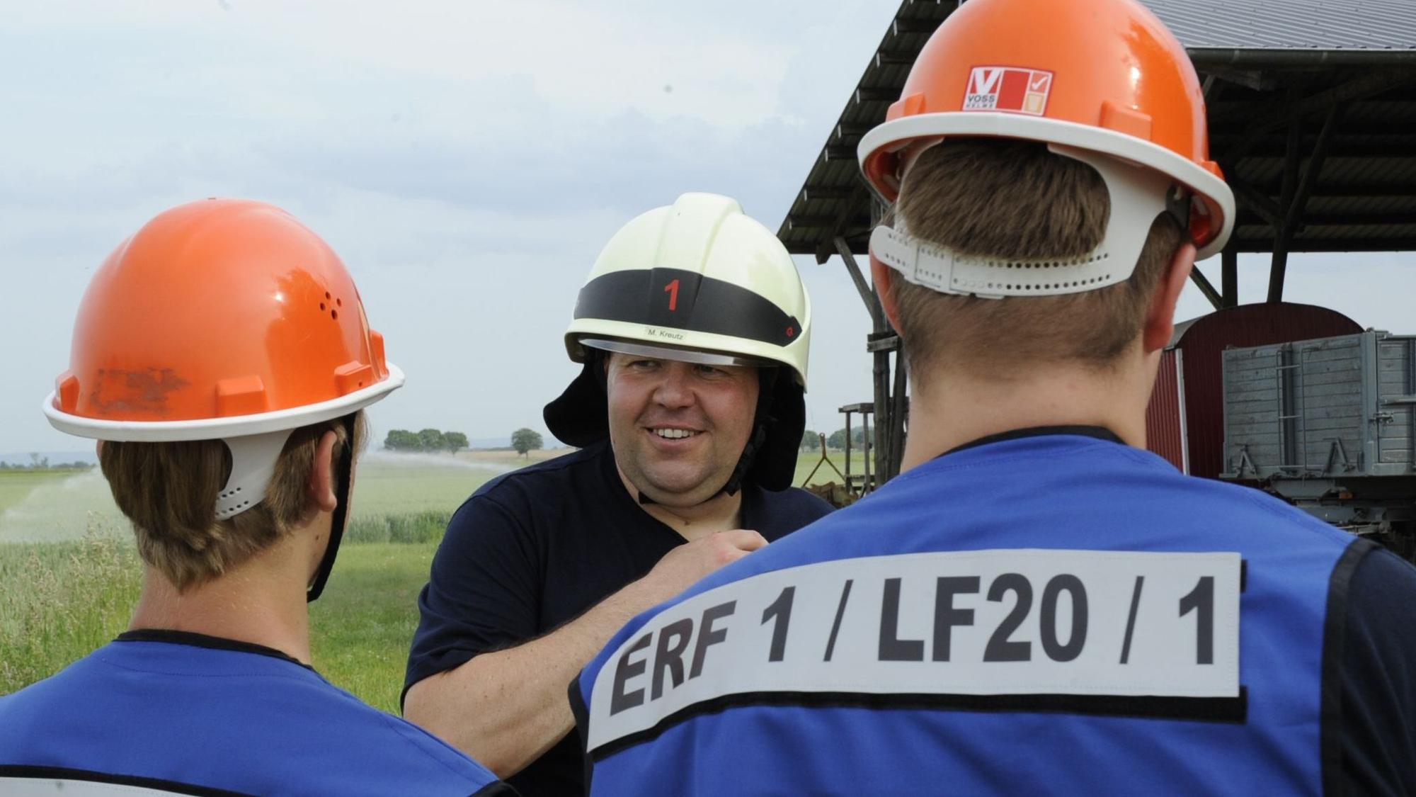 Ein Mann mit Helm spricht mit zwei anderen jungen Männern in orangenen Helmen, die von hinten zu sehen sind.