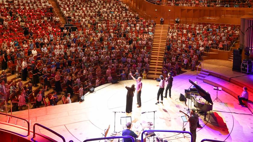 1.000 Kölner Grundschülerinnen und -schüler sitzen in den Rängen der Kölner Philharmonie und singen, auf der Bühne stehen vier Singleiterinnen.