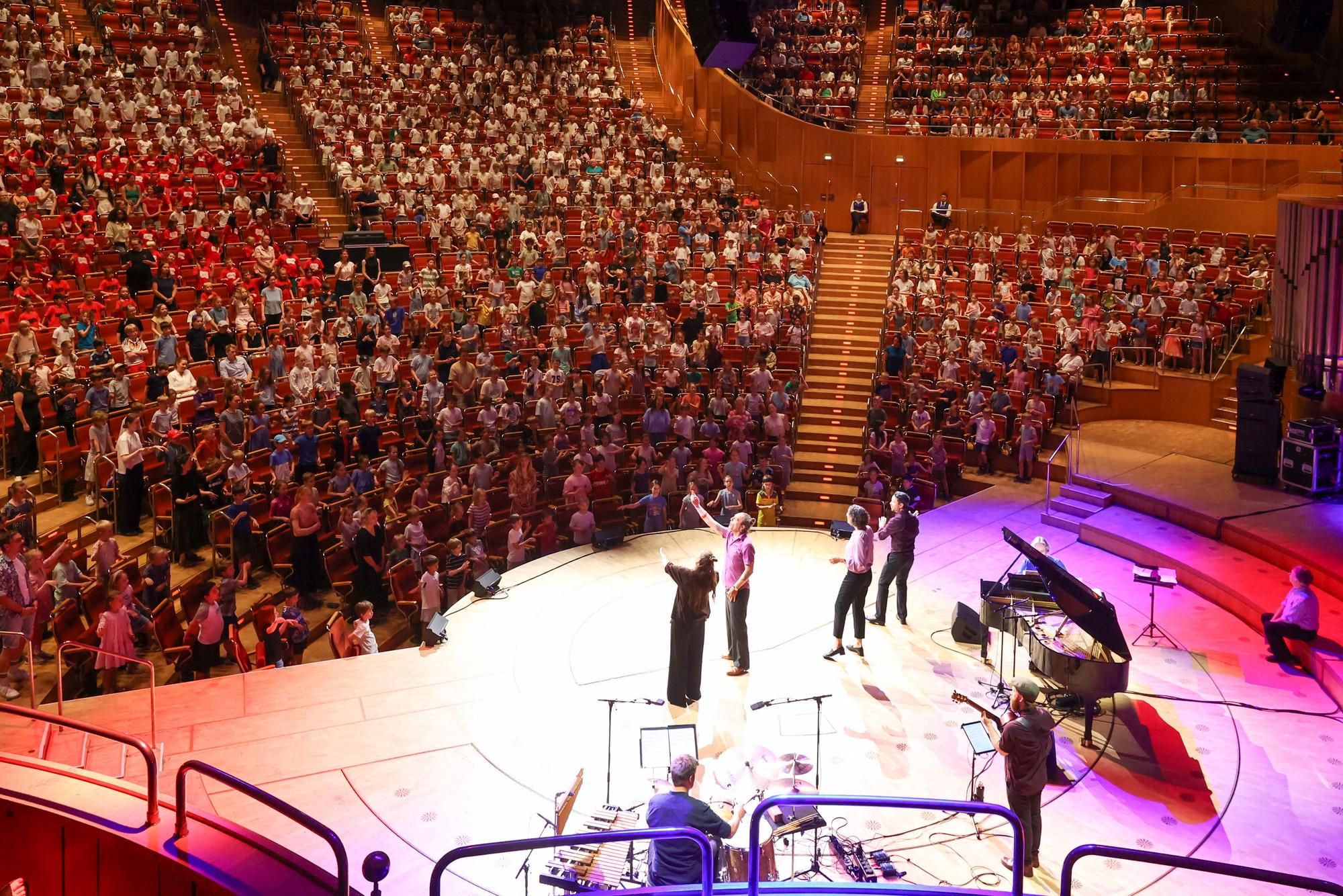 1.000 Kölner Grundschülerinnen und -schüler sitzen in den Rängen der Kölner Philharmonie und singen, auf der Bühne stehen vier Singleiterinnen.