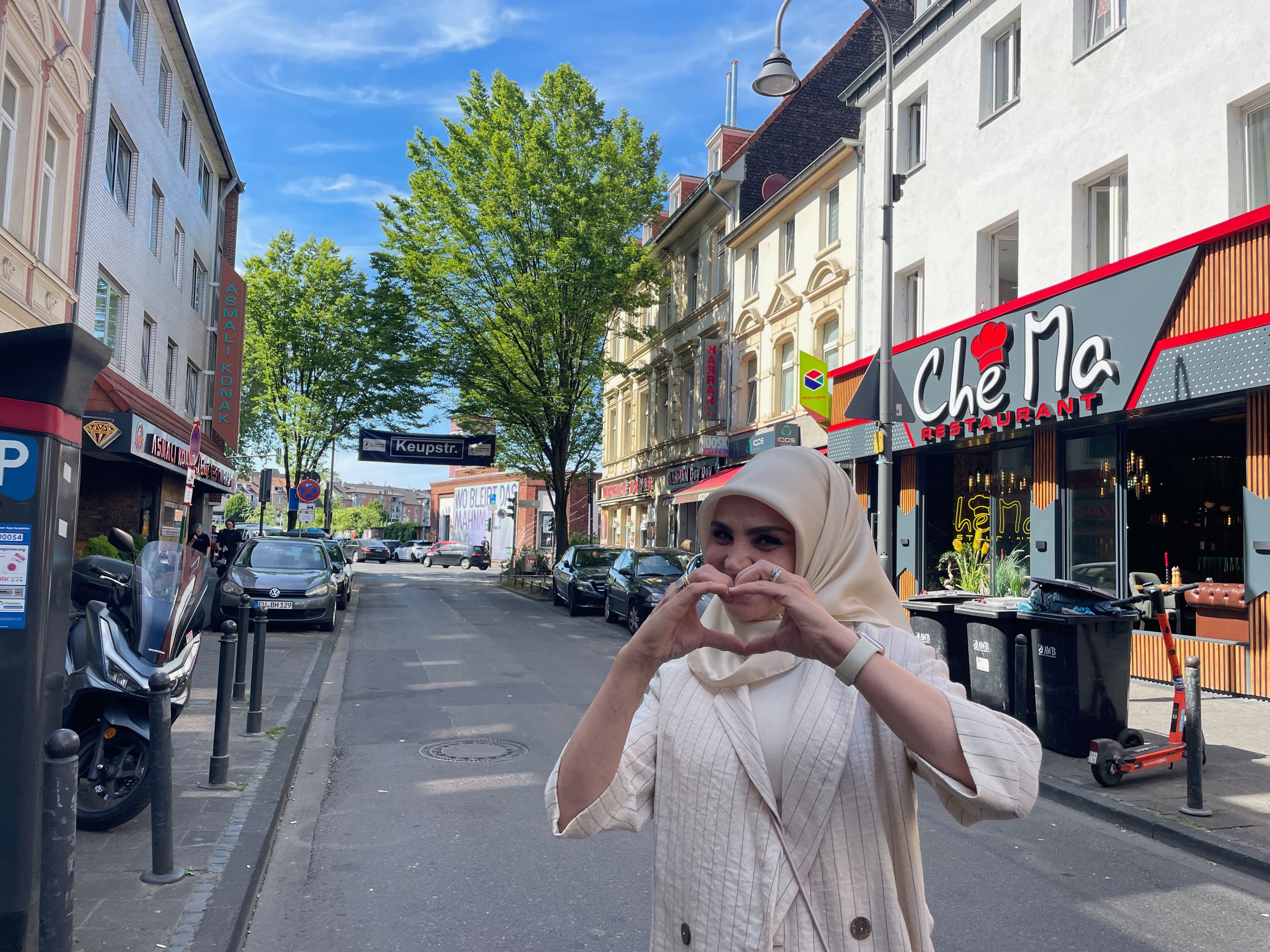 Eine Frau mit hellem Gewand und Kopftuch steht auf einer schmalen Straße und formt mit den Händen ein Herz.