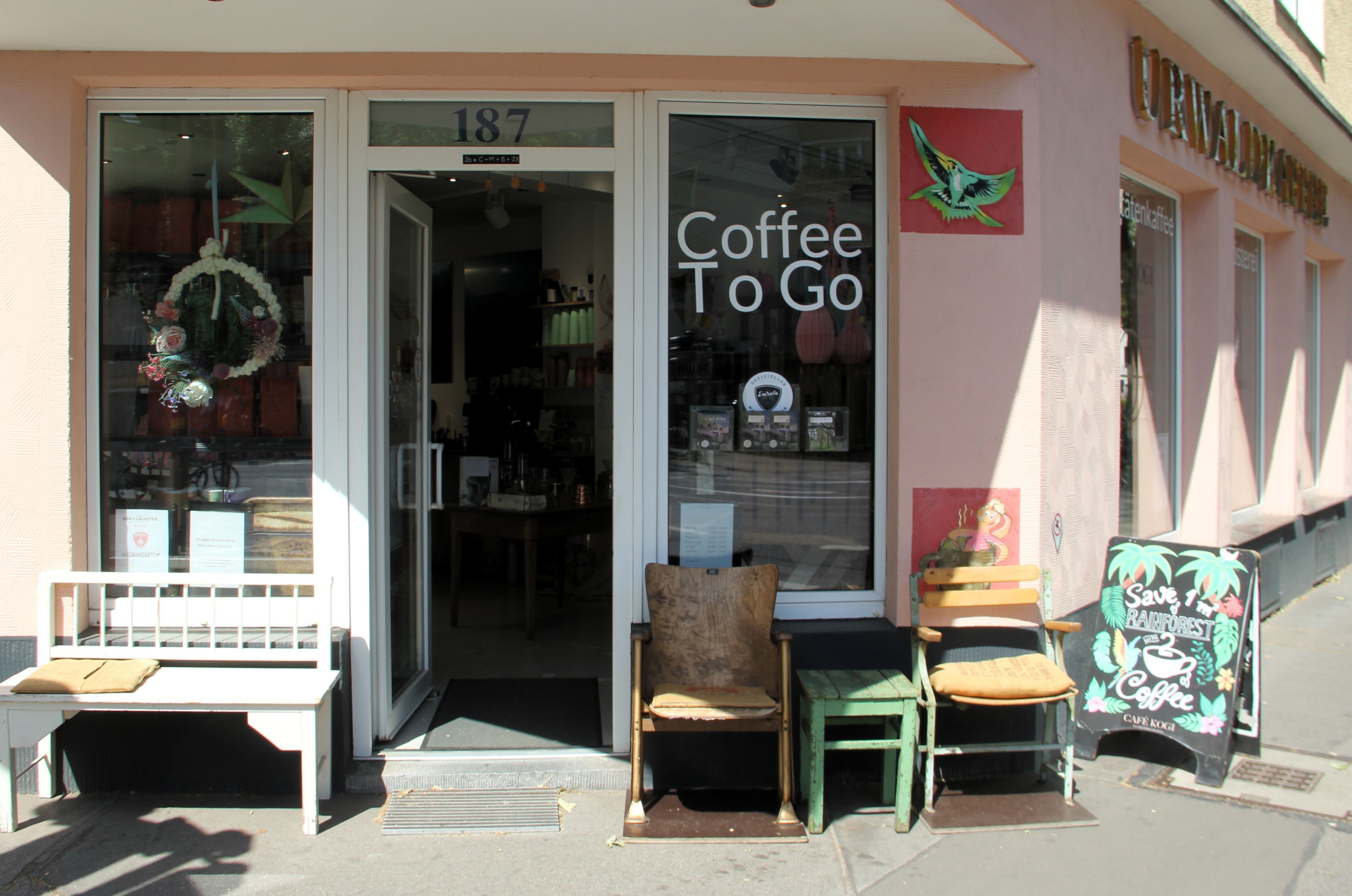 Die Front eines Cafés ist zu sehen. Es stehen Bänke und Stühle davor, an der Seite ist der Schriftzug Urwaldkaffee angebracht.