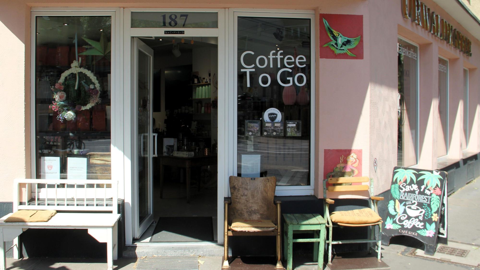 Die Front eines Cafés ist zu sehen. Es stehen Bänke und Stühle davor, an der Seite ist der Schriftzug Urwaldkaffee angebracht.