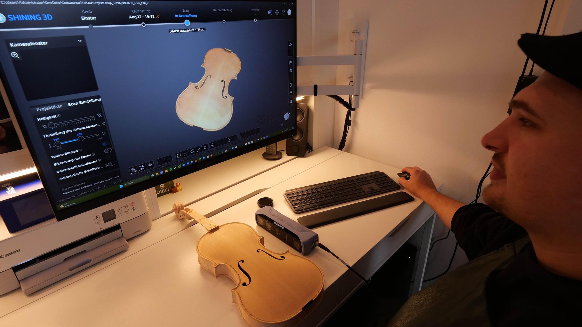 Ein Mann mit Kappe sitzt vor einem Monitor, auf dem der Korpus einer Geige zu sehen ist. Eine echte Geige liegt vor ihm auf dem Schreibtisch.