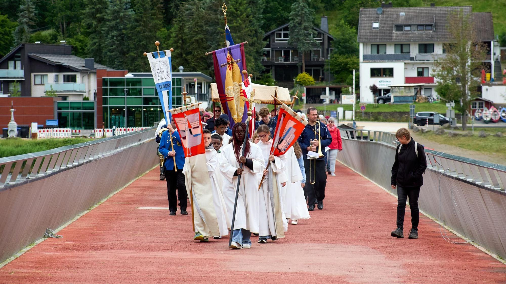 Ein Gruppe Messdienerinnen und Messdiener zieht mit Fahnen vor einer Menschengruppe über eine Brücke.