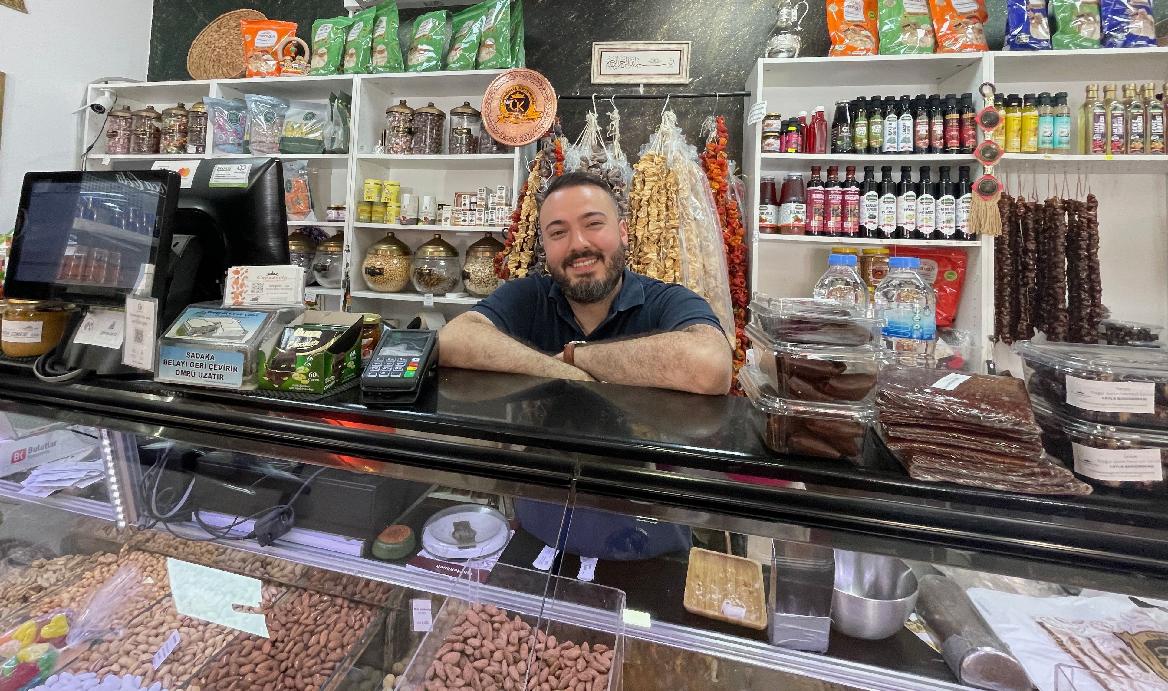 Ein junger Mann mit dunklem Haar und Bart steht hinter einer Frischetheke in einem Laden.