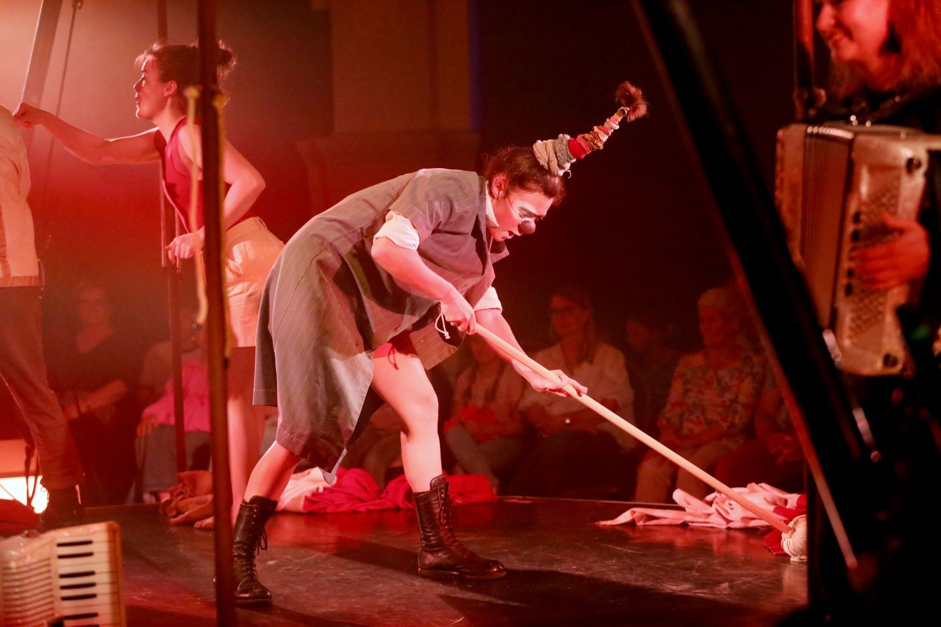Eien Frau im Clownskostüm und mit roter Nase hantiert mit einem Wischmopp auf der Bühne.