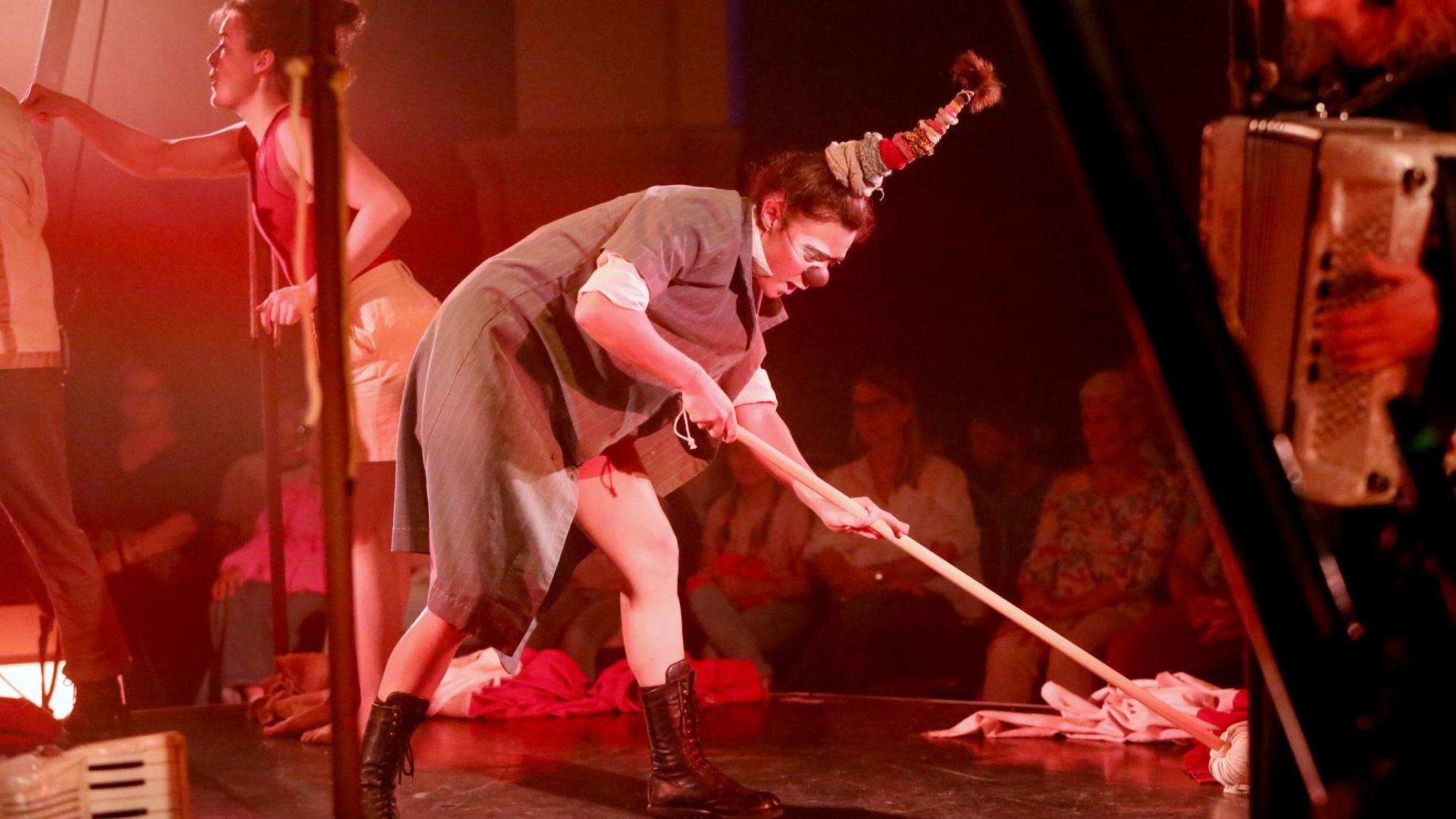 Eien Frau im Clownskostüm und mit roter Nase hantiert mit einem Wischmopp auf der Bühne.