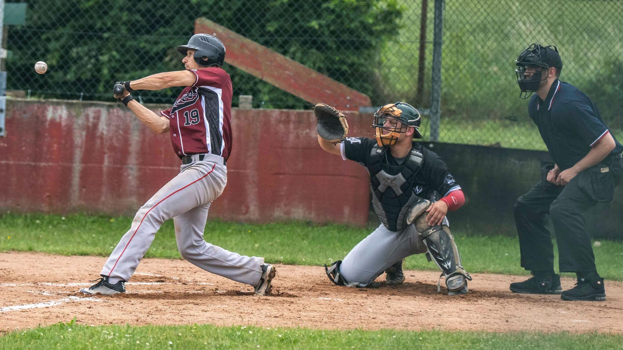 Ein Baseballspieler beim Schlag. Die Muskeln sind angespannt. Hinter ihm der Catcher und der Umpire.