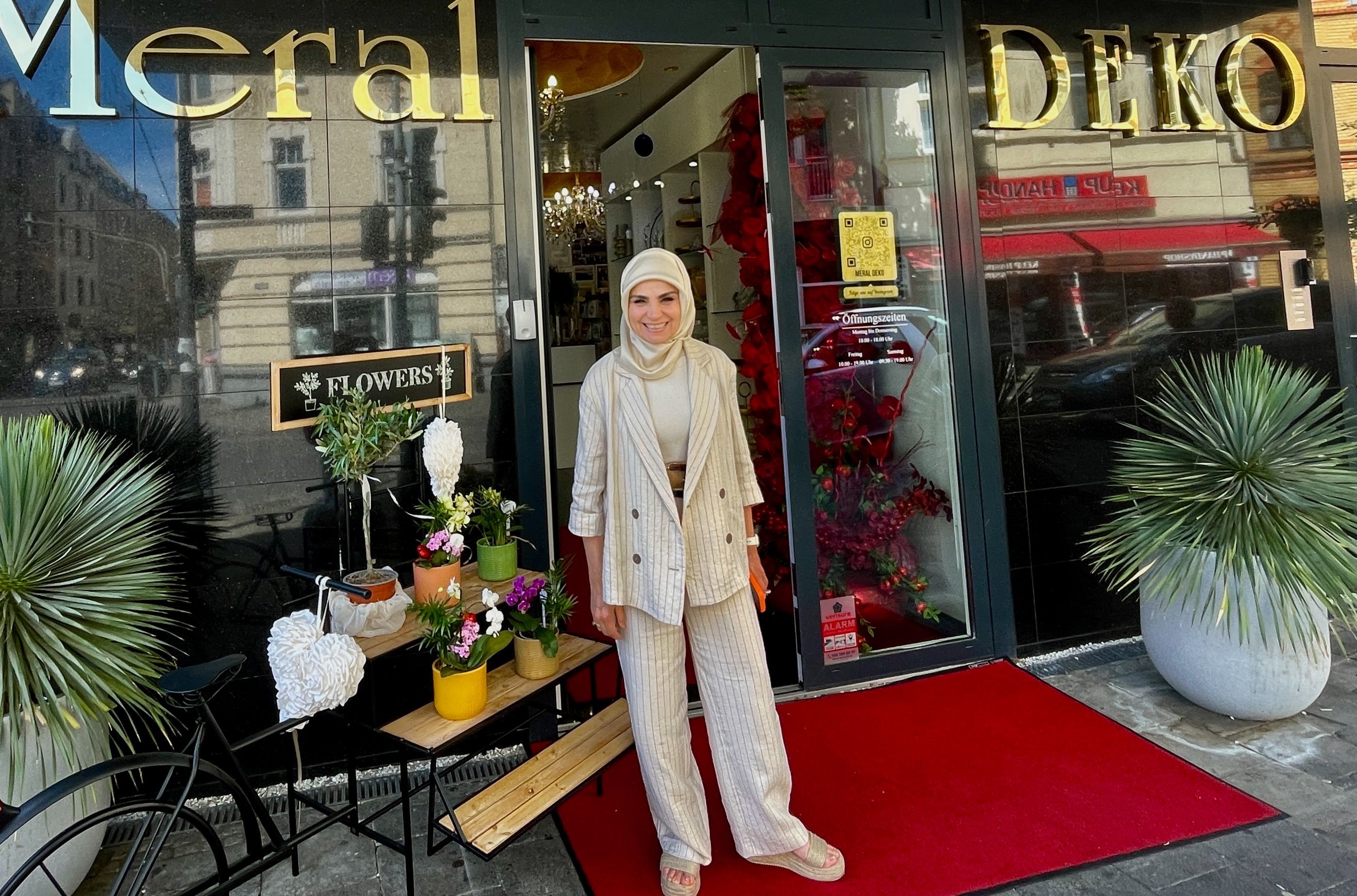 Eine Frau in hellem Blazer, heller Hose und hellem Kopftuch steht vor dem Schaufenster ihres Ladens.