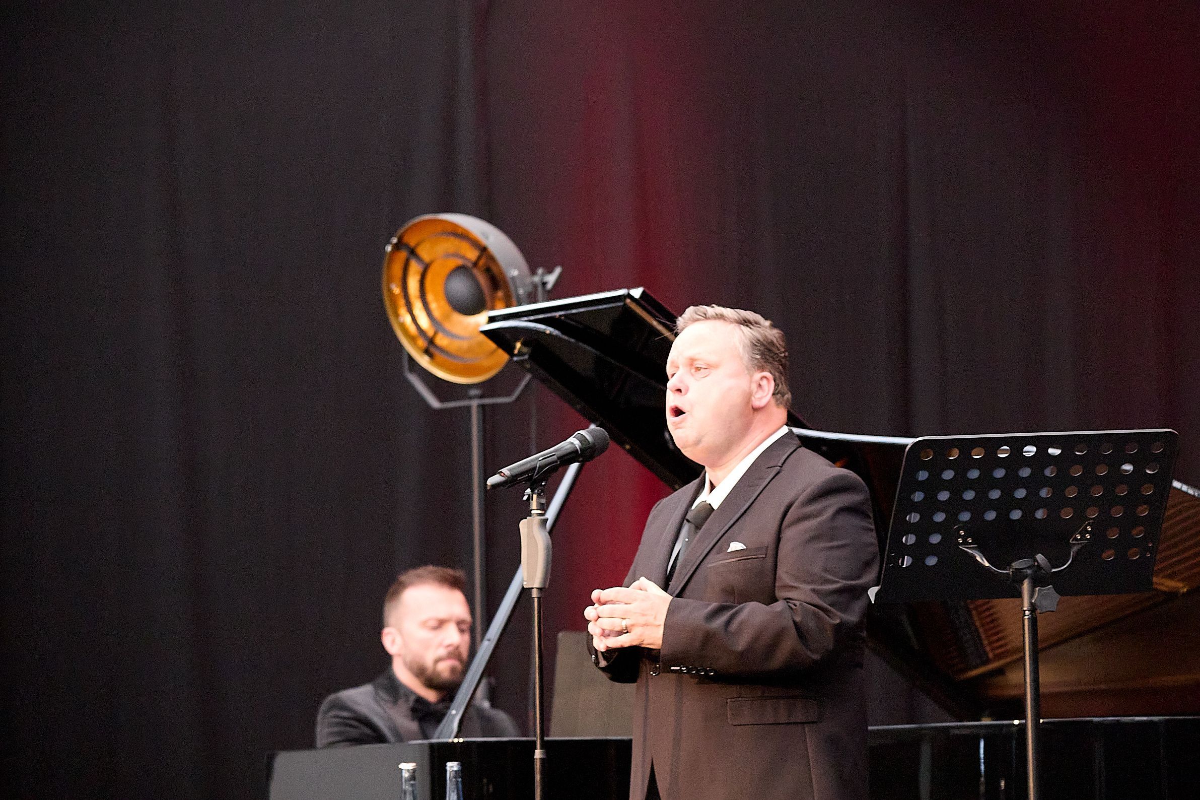 Der Tenor Paul Potts singt in ein Mikrofon, im Hintergrund ist Vytis Šakūras am Piano zu sehen.