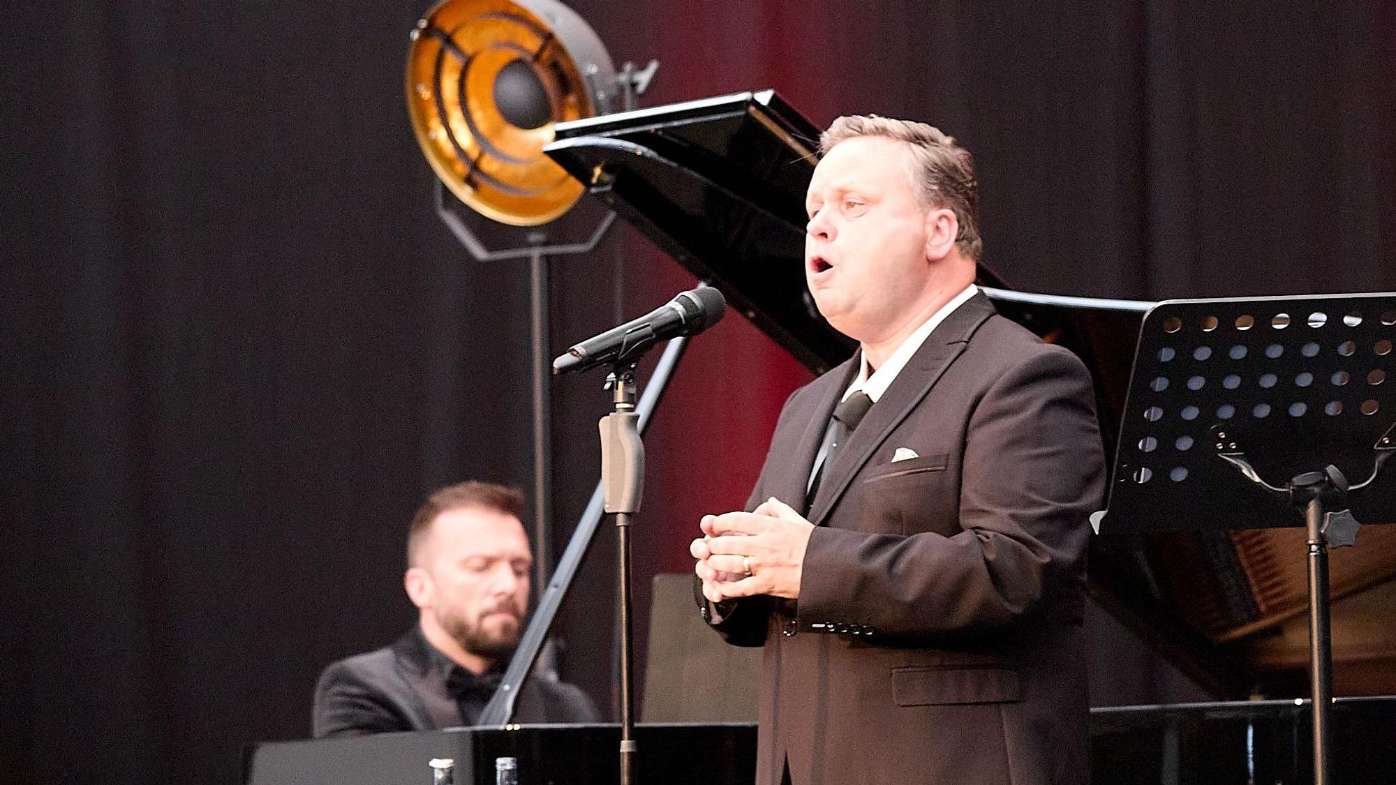 Der Tenor Paul Potts singt in ein Mikrofon, im Hintergrund ist Vytis Šakūras am Piano zu sehen.