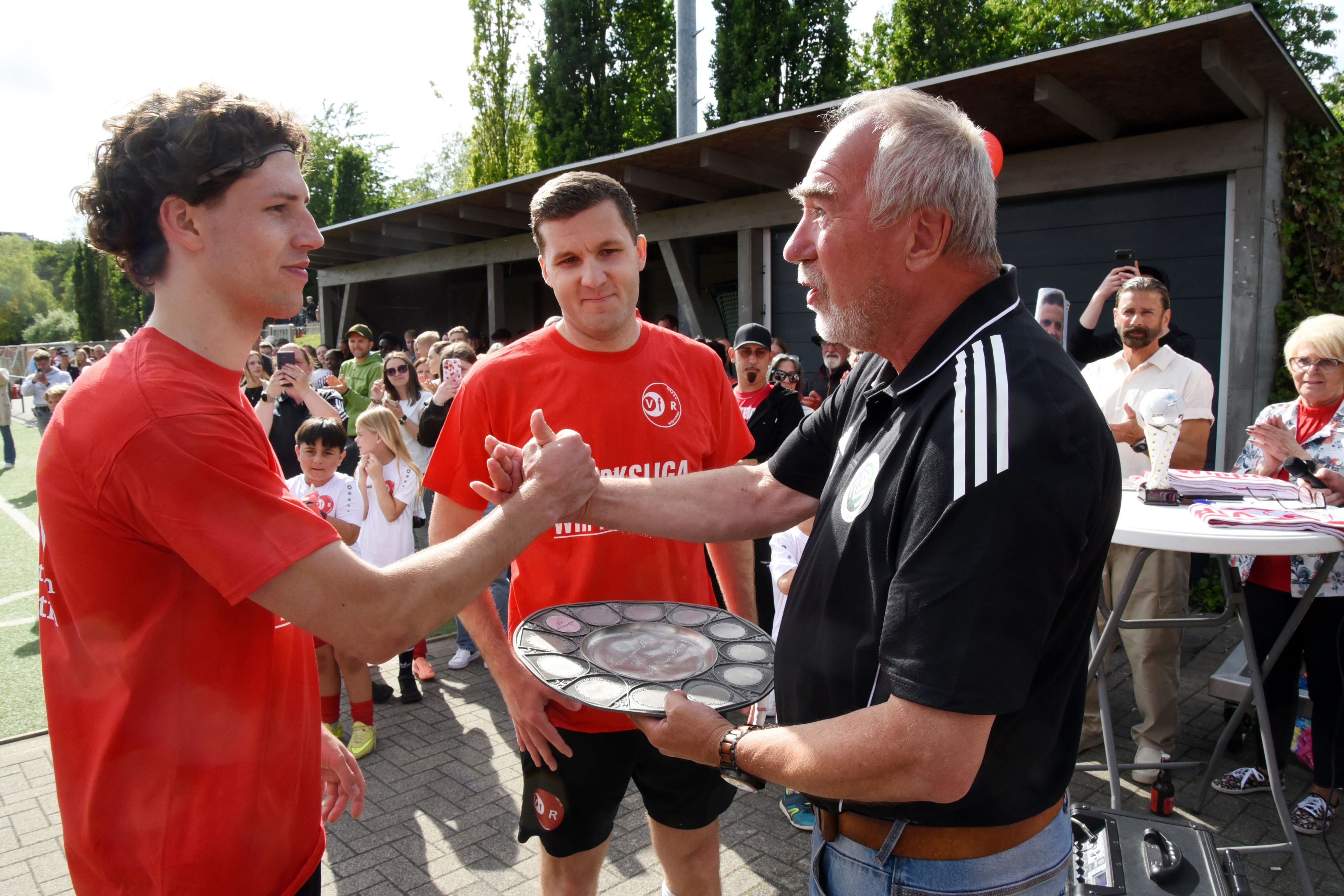 Zu sehen ist die Übergabe einer Meisterschale durch einen Sportfunktionär an zwei Fußballspieler.