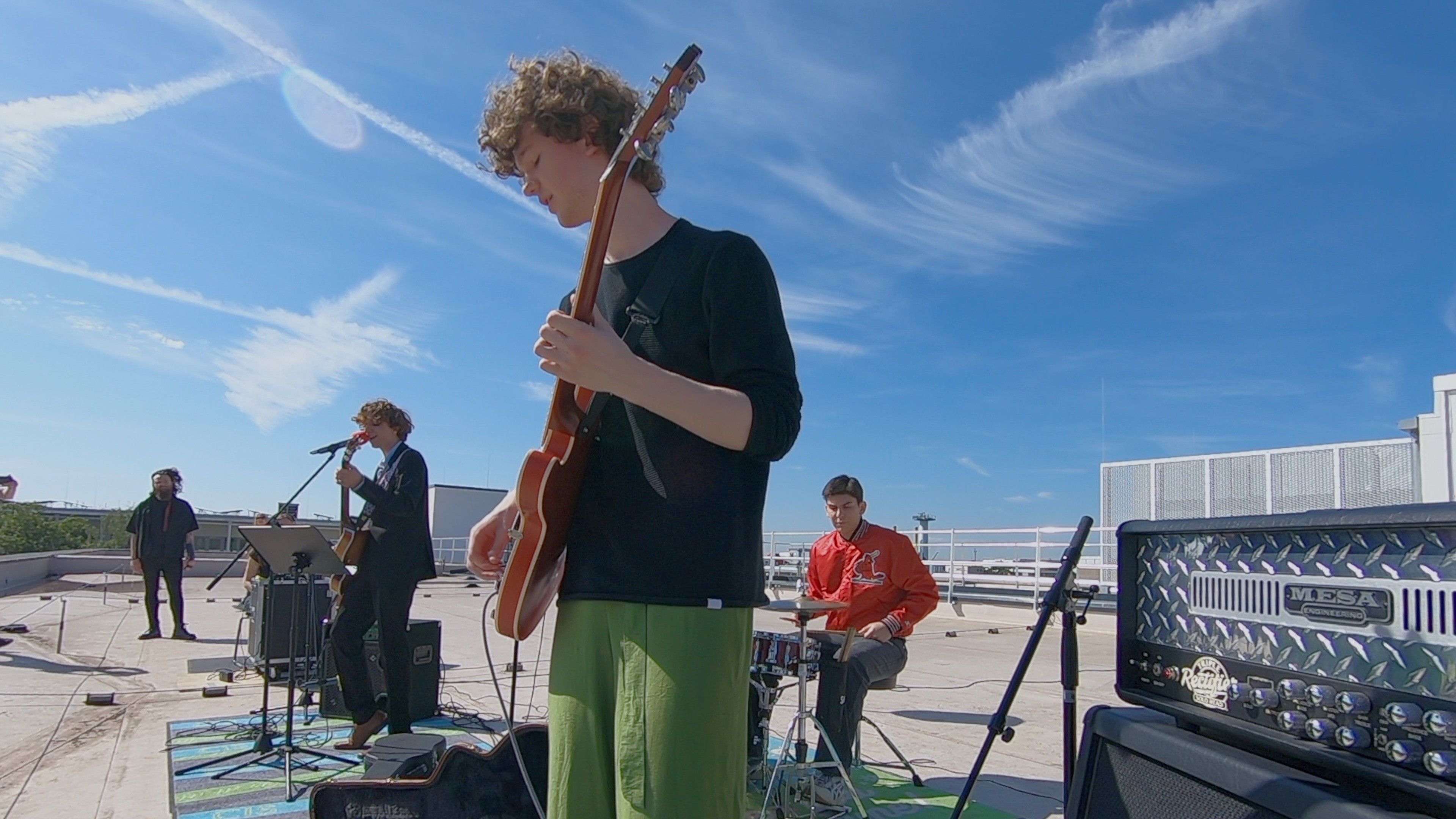 Ein Mann und drei junge Männer an Instrumenten sind auf einem Flachdach vor strahlend blauem Himmel.