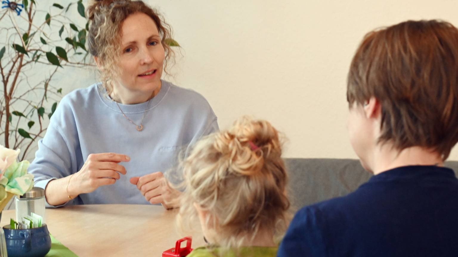 Eine Frau mit Locken sitzt einer Mutter gegenüber, die ein blondgelocktes Kind auf dem Schoß hat.