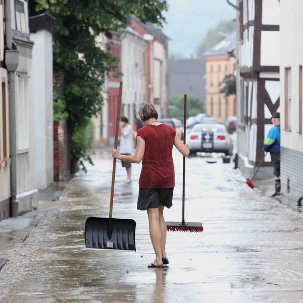 Eine Frau mit Besen und Schaufel auf einer von braunem Wasser überfluteten Straße.
