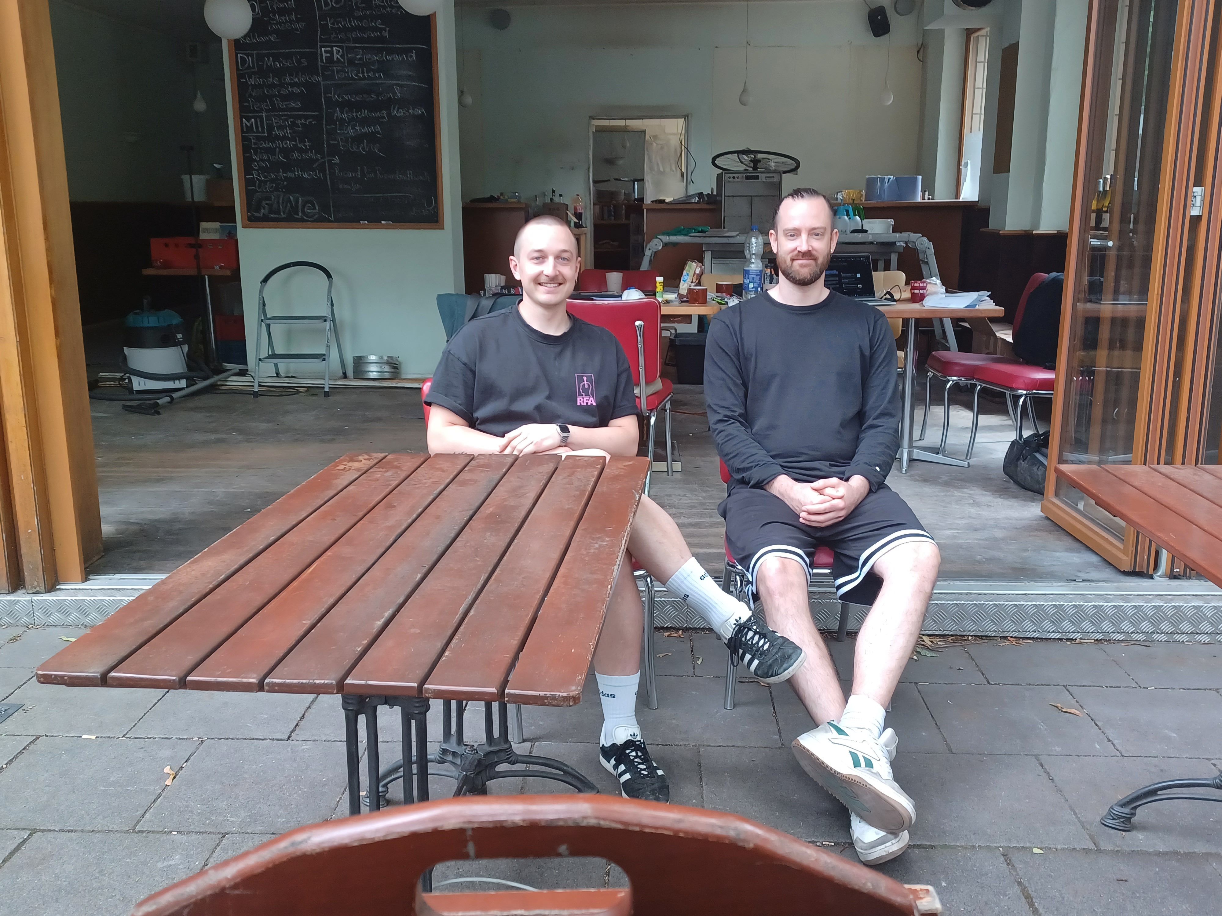 Zwei junge Männer sitzen auf einer Terrasse vor einem Café, das gerade umgebaut wird.