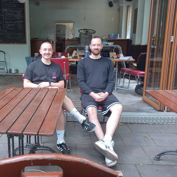 Zwei junge Männer sitzen auf einer Terrasse vor einem Café, das gerade umgebaut wird.