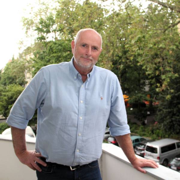 Ein Mann mit kurzen grauen Haaren und blauem Hemd steht auf einem Balkon, hinter ihm sieht man einen Park und parkende Fahrzeuge.