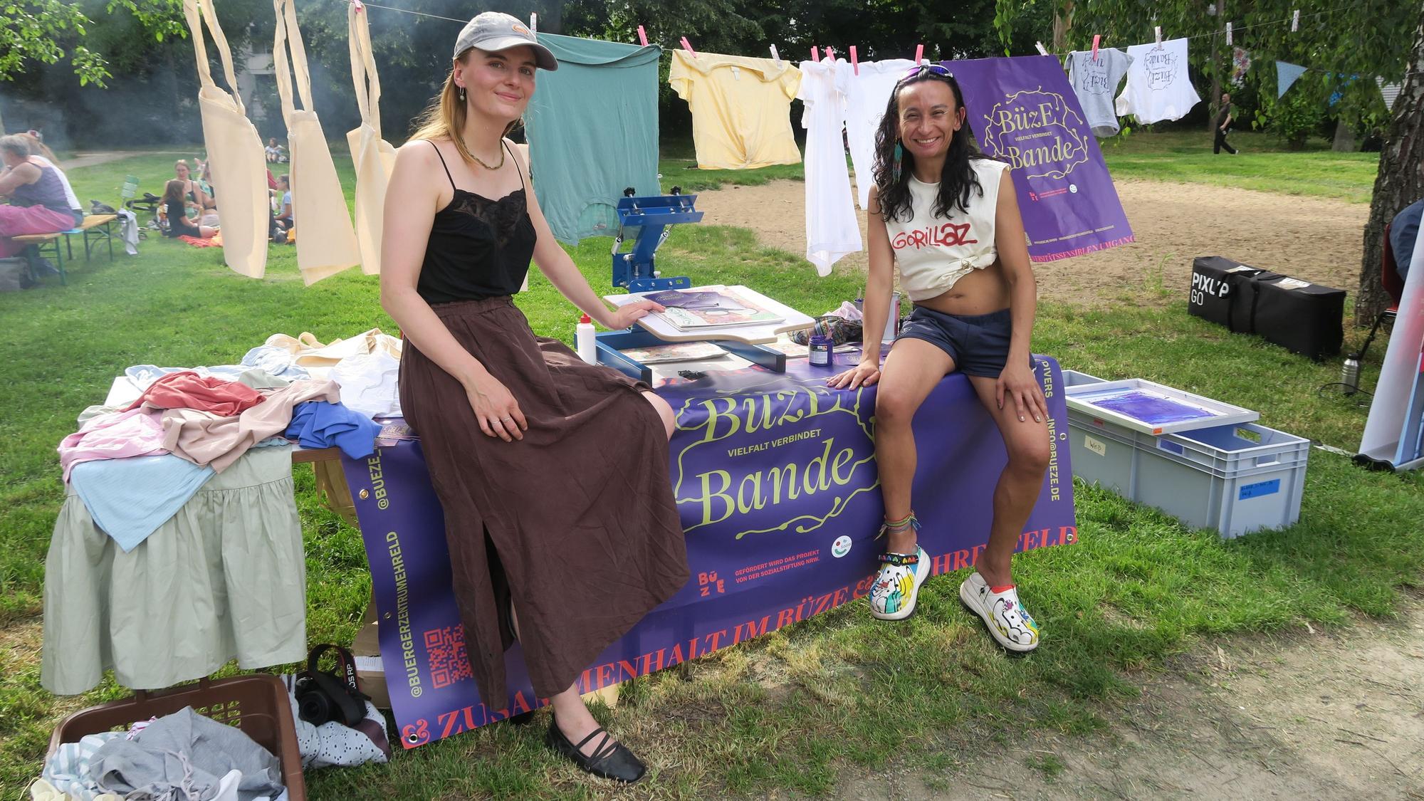 Zwei Frauen sitzen auf einem Tisch, auf dem Textiliien und Info-Material liegen.