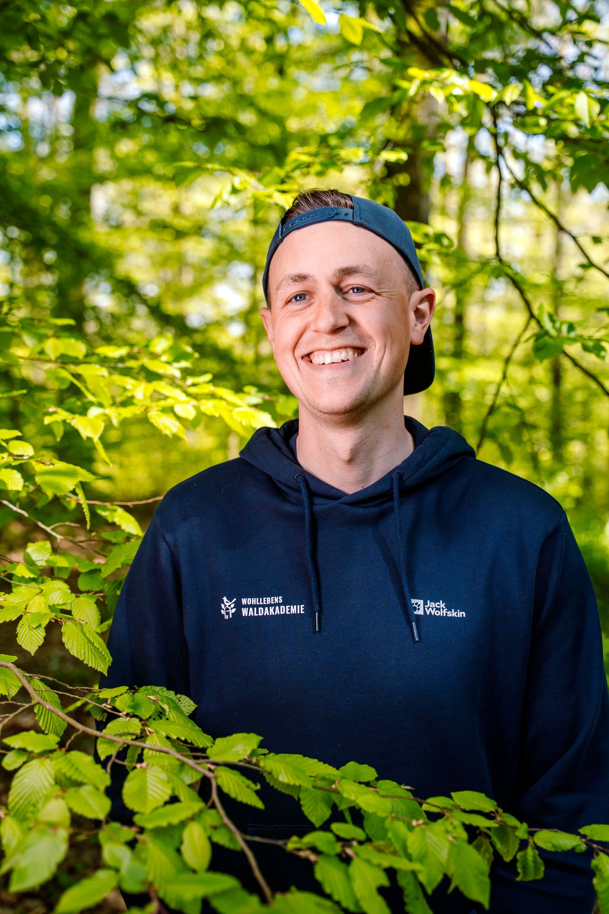 Tobias Wohlleben steht im Wald, er trägt eine Kappe verkehrt herum.