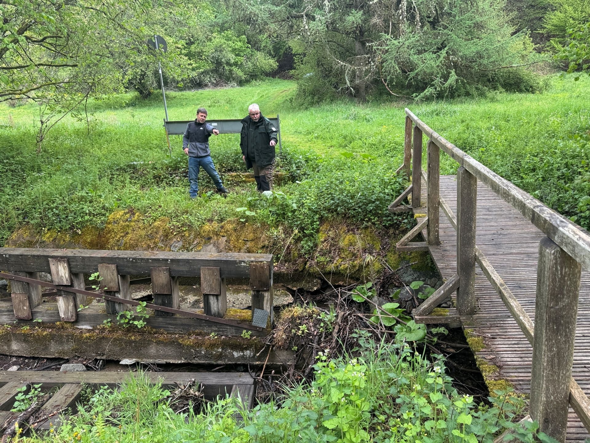 Zwei Männer stehen am Ufer und schauen auf eine beschädigte Holzbrücke.
