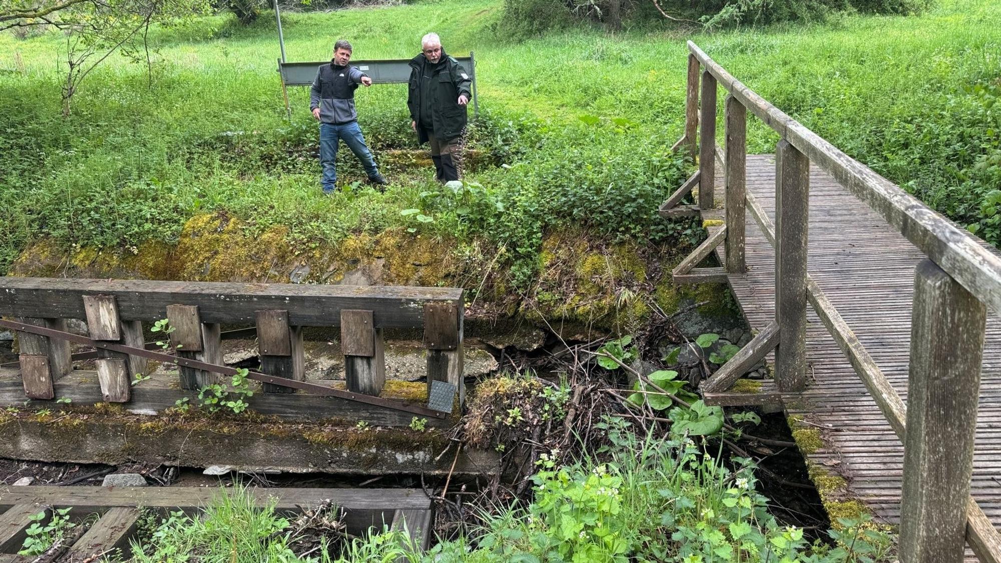 Zwei Männer stehen am Ufer und schauen auf eine beschädigte Holzbrücke.