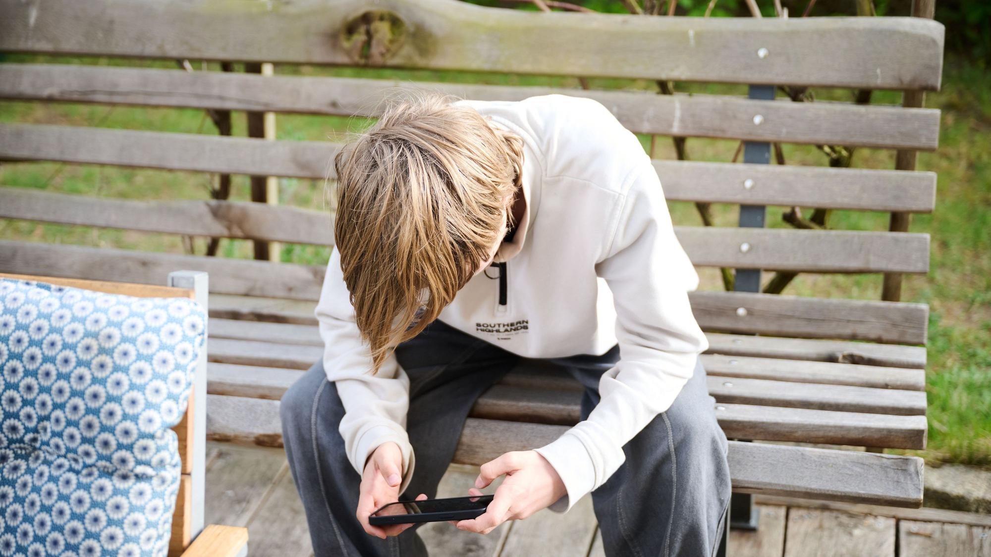 Zu sehen ist ein Jugendlicher auf einer Bank, der auf sein Handy schaut.