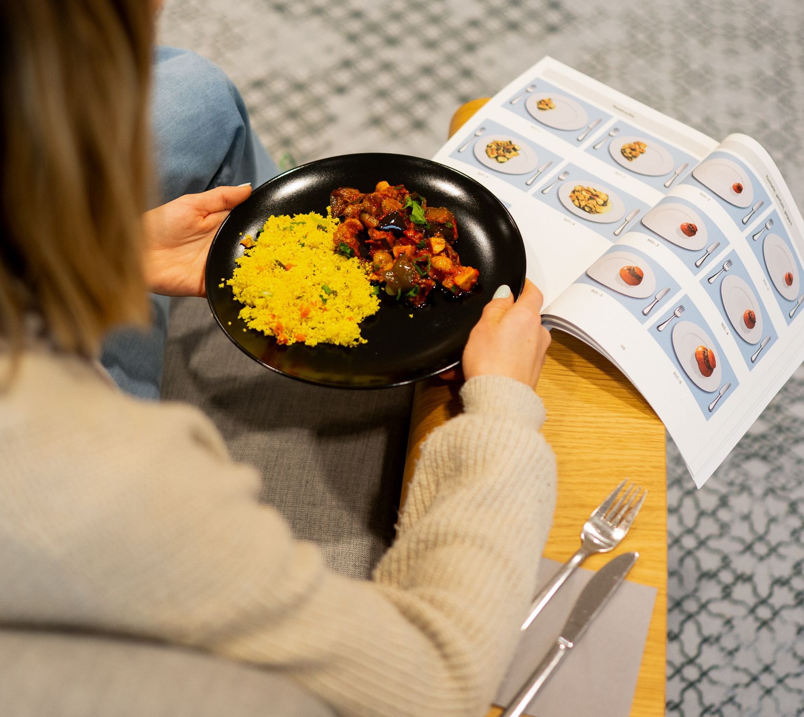 Das Foto zeigt eine Frau von hinten, die einen Teller mit einer Speise in den Händen hält, davor ein Buch mit Abbildungen von Speise-Portionsgrößen.