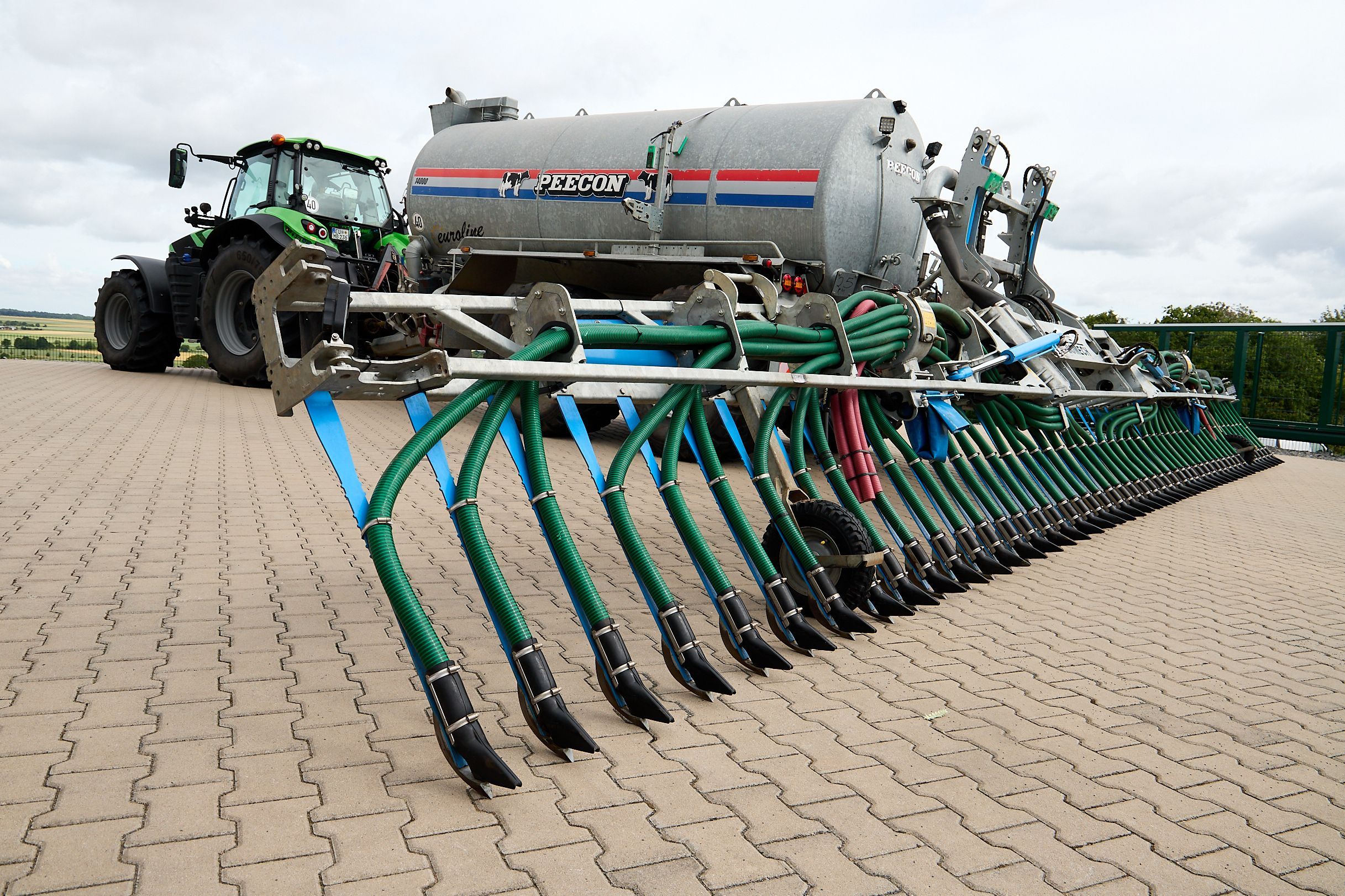 Ein Traktor steht auf einer gepflasterten Fläche. Er zieht ein großes Fass mit einer Vorrichtung, die Gülle dicht am Boden ausbringt.