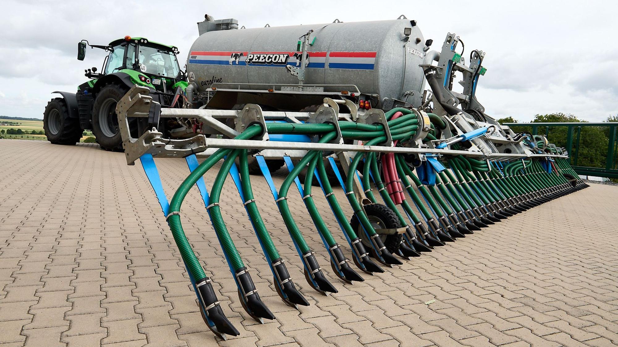 Ein Traktor steht auf einer gepflasterten Fläche. Er zieht ein großes Fass mit einer Vorrichtung, die Gülle dicht am Boden ausbringt.