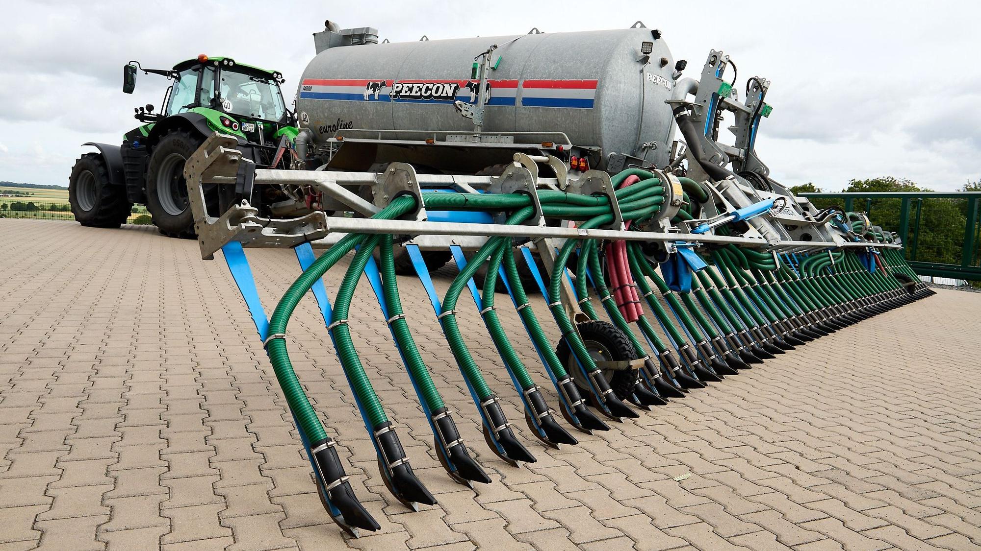 Ein Traktor mit einem Schleppschuhverteiler, mit dem Gülle bodennah ausgebracht werden kann, steht auf einem gepflasterten Hof.